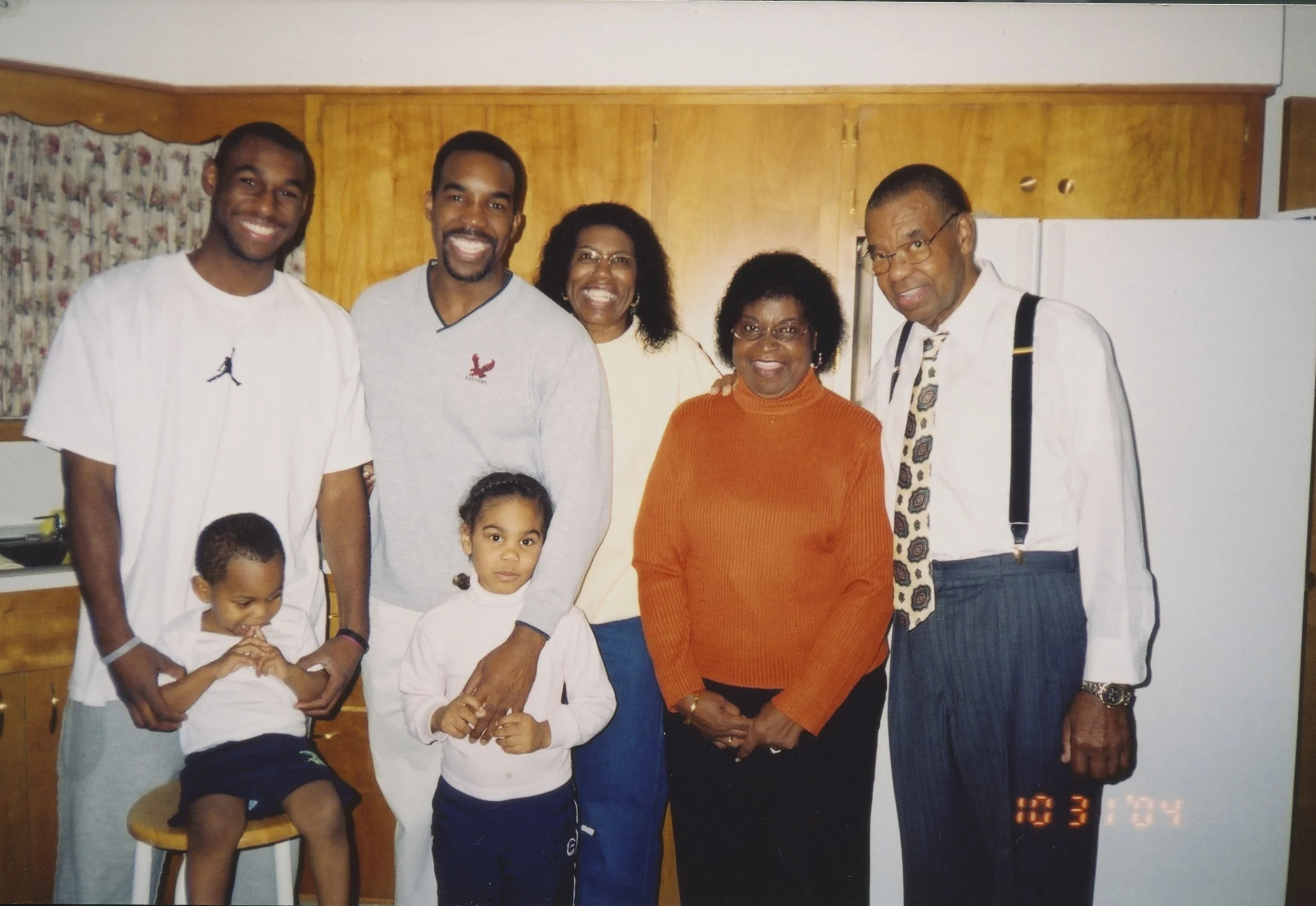 Family group photo in a kitchen, featuring adults and children, some smiling and some with neutral expressions.