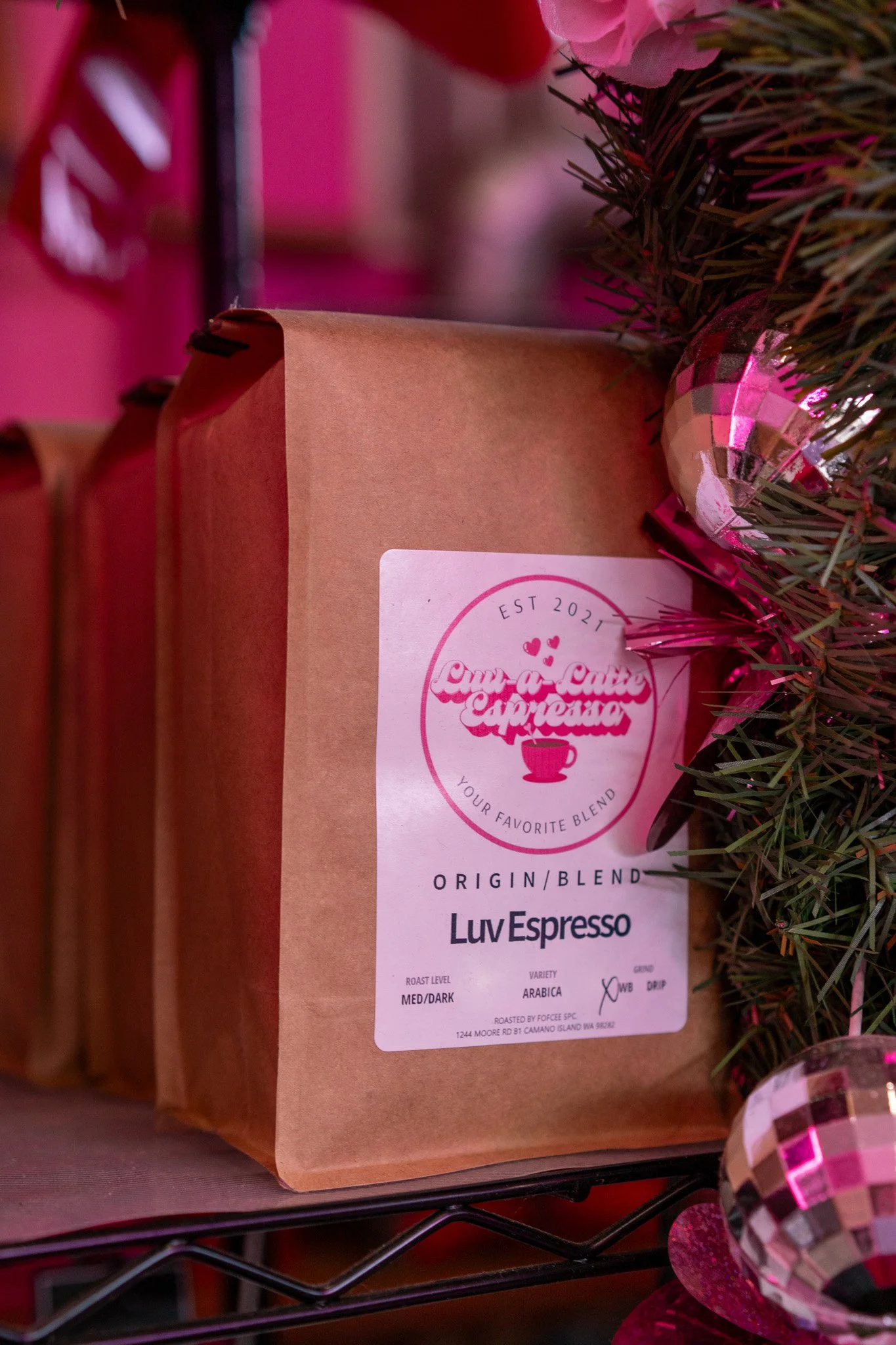 Brown paper bag of coffee labeled LuvEspresso on a shelf decorated with pink and silver Christmas ornaments.
