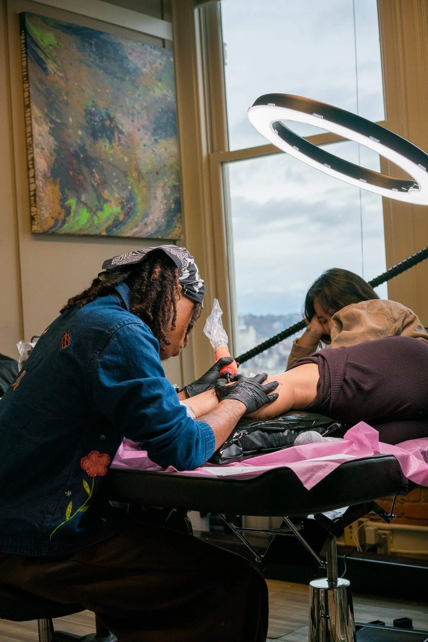 Tattoo artist working on a client's leg in art studio with large window and colorful painting on wall.