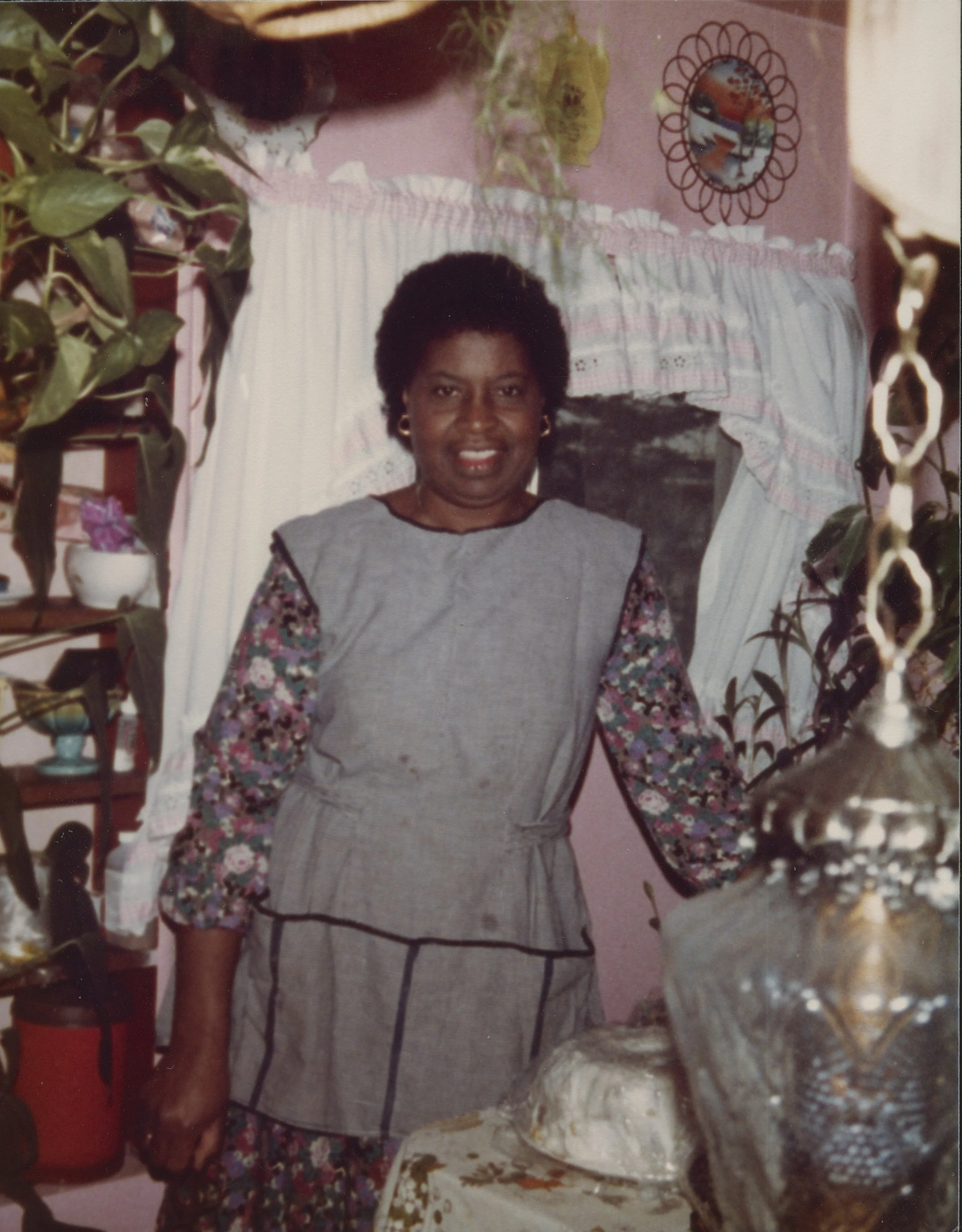 A woman standing indoors in front of a pink wall decorated with a mirror and plants, smiling at the camera.