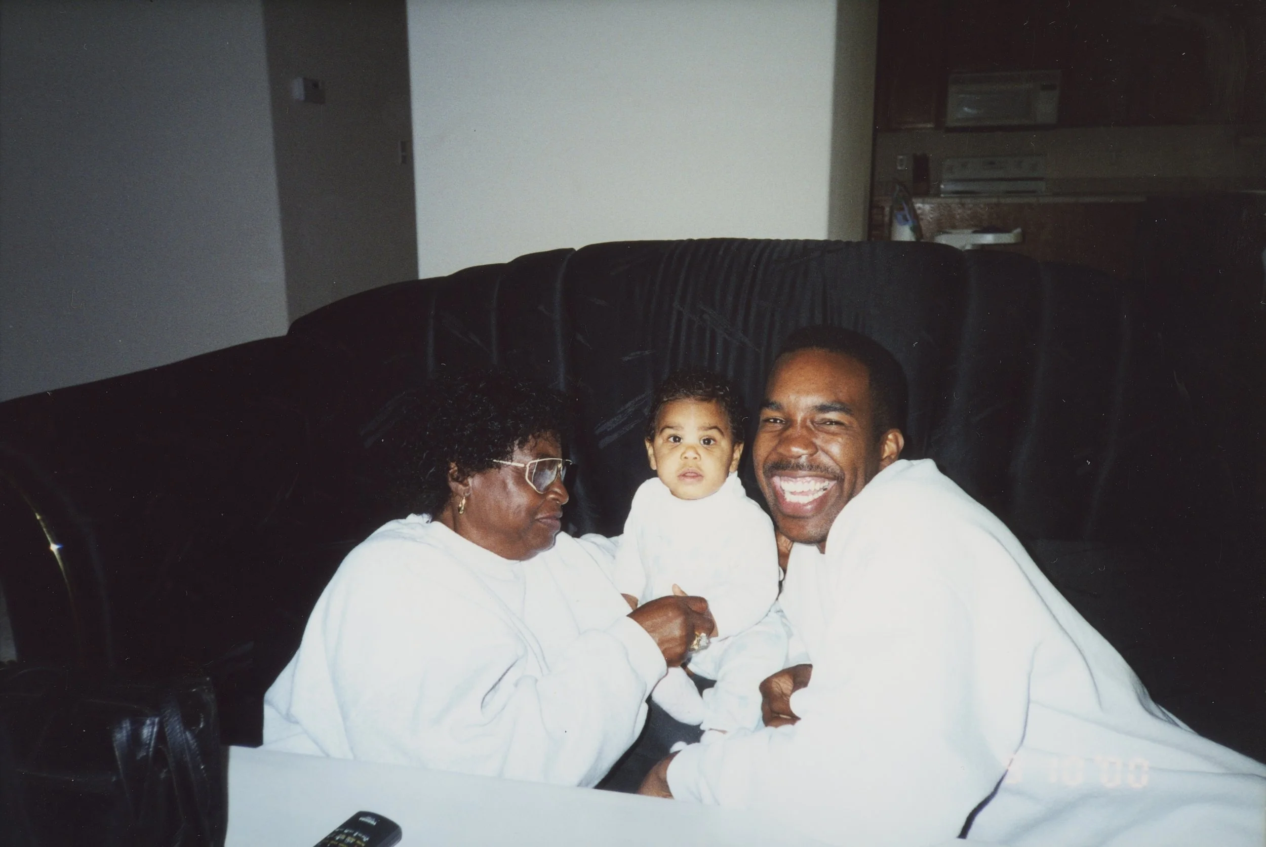 A woman, a man, and a young child sitting on a black couch inside a home. The woman is wearing glasses and white clothing, looking at the child. The man is smiling widely, also in white clothing, facing the camera. In the background, a kitchen with a