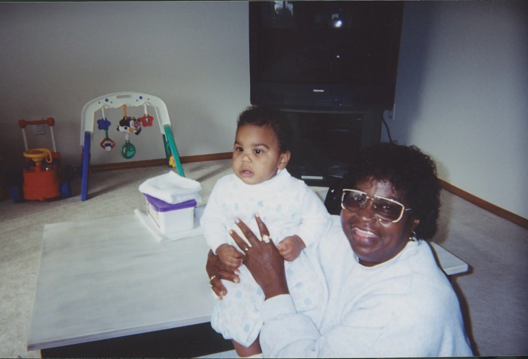 A woman with large glasses and curly hair holding a young child in a room with a TV, a small table, and toys in the background.