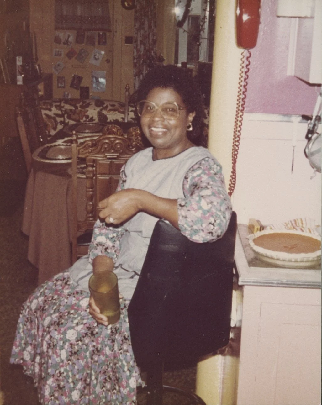 A woman with glasses, smiling, sitting in a vintage kitchen with a floral dress and holding a glass. A pie is on the counter nearby. The background shows a wooden dining area with a table, chairs, and a wall decorated with photographs.