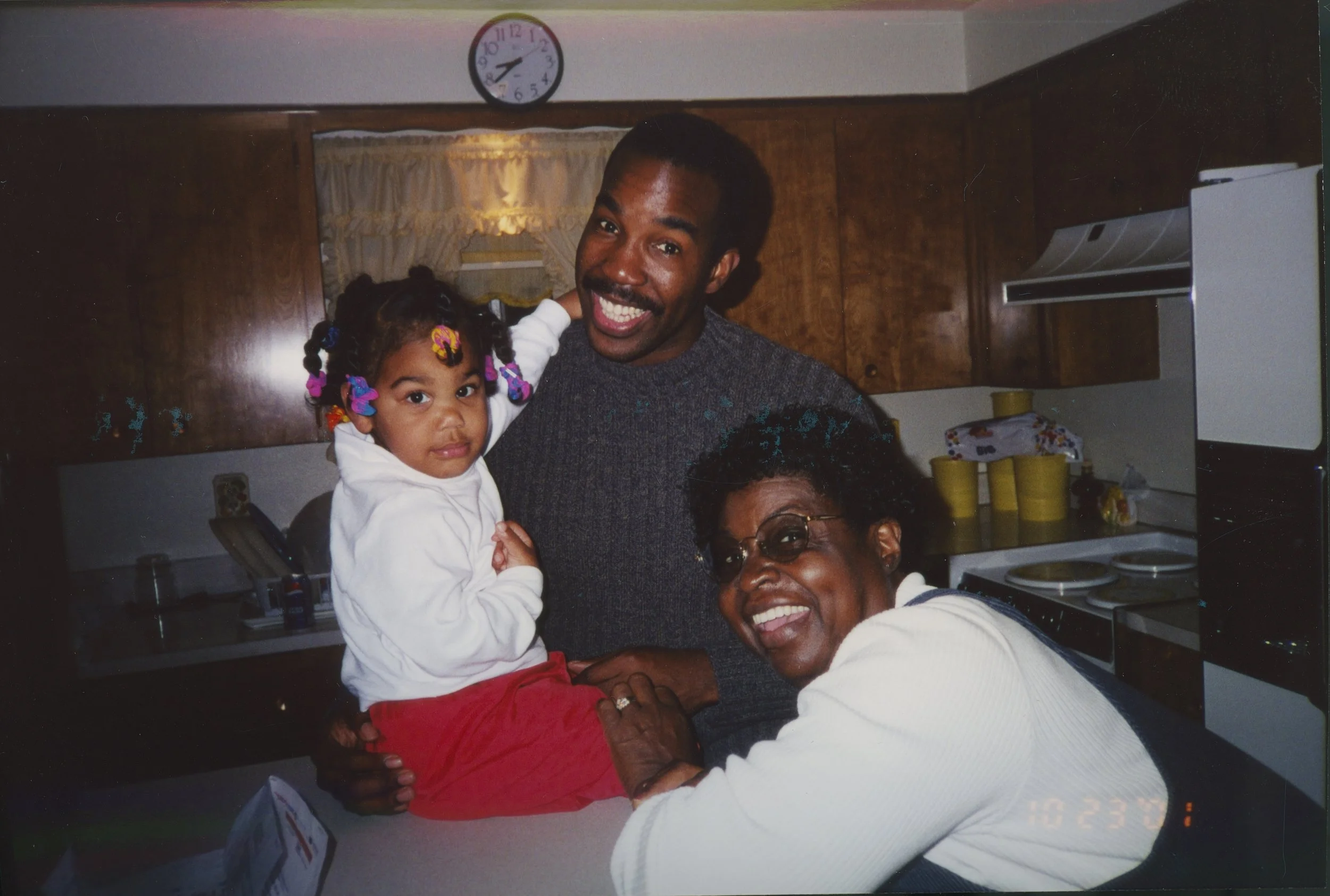 A young girl with colorful braids sitting on a kitchen counter with an older woman and a man smiling nearby.