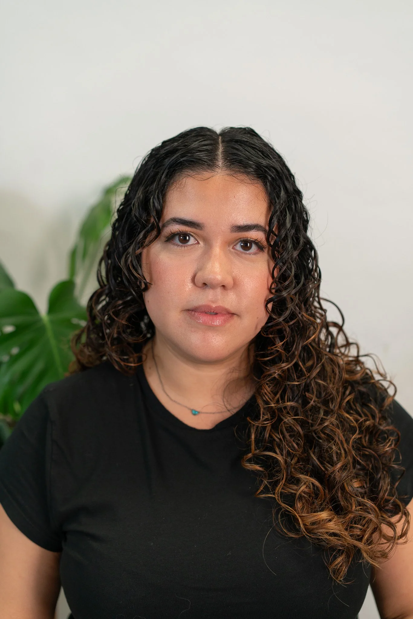 A woman with dark, curly hair and a neutral expression, wearing a black shirt and a necklace, standing in front of a white wall and green plant.
