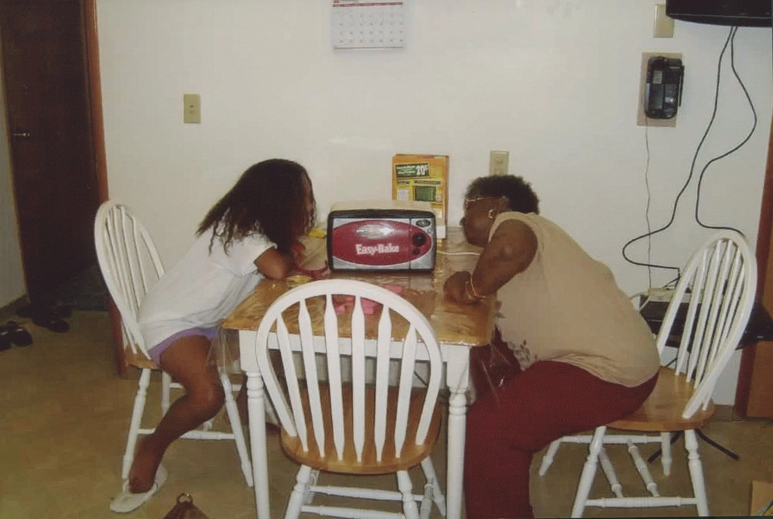 A young girl and an older woman are leaning on a wooden dining table with a red and gray EasyBake oven in the center. The girl is wearing a white top and shorts, and the woman is in a beige top and red pants. They are facing each other, with the girl