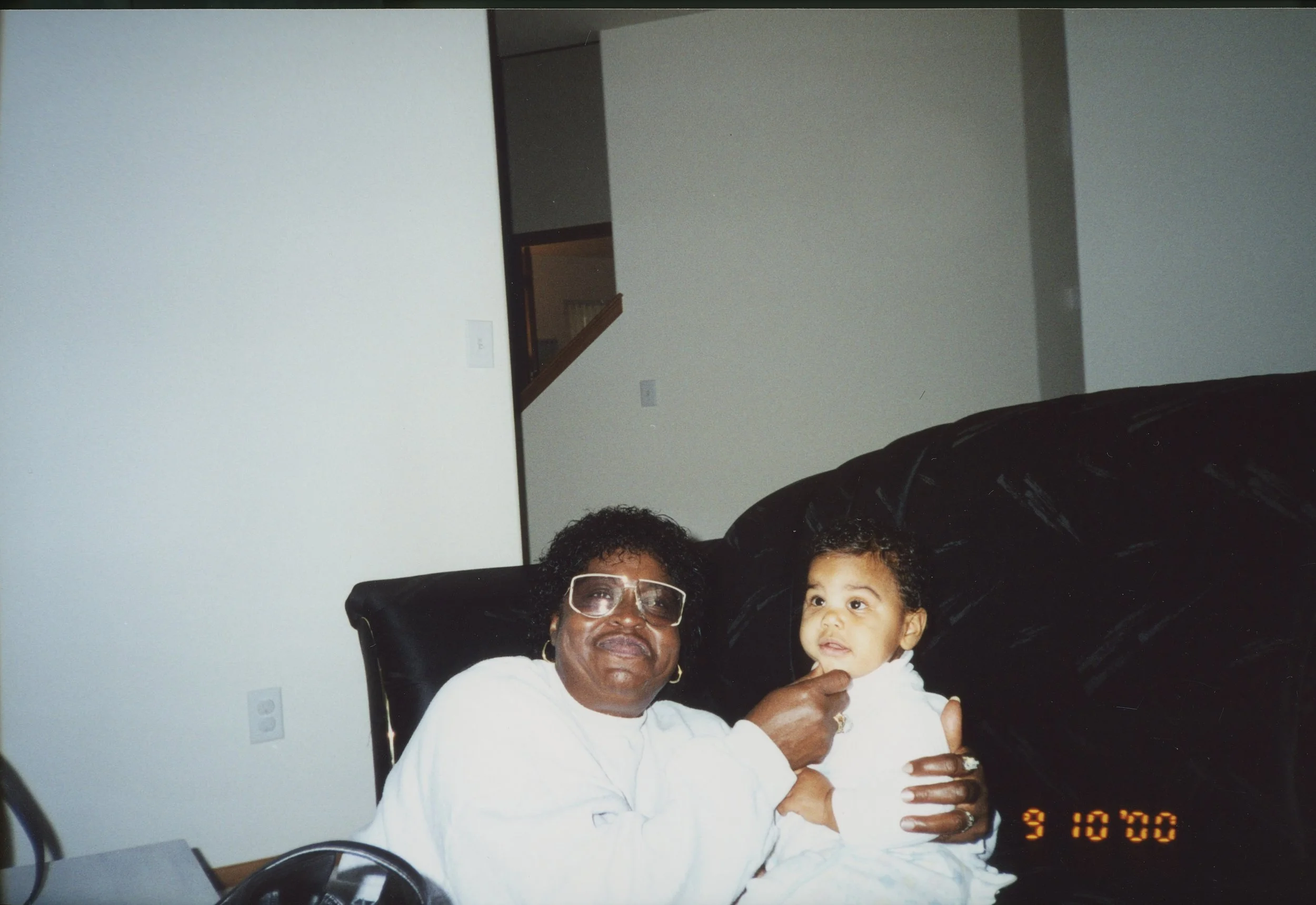 Older woman with glasses and earrings holding a young girl dressed in white, sitting in a living room with beige walls and a black couch.