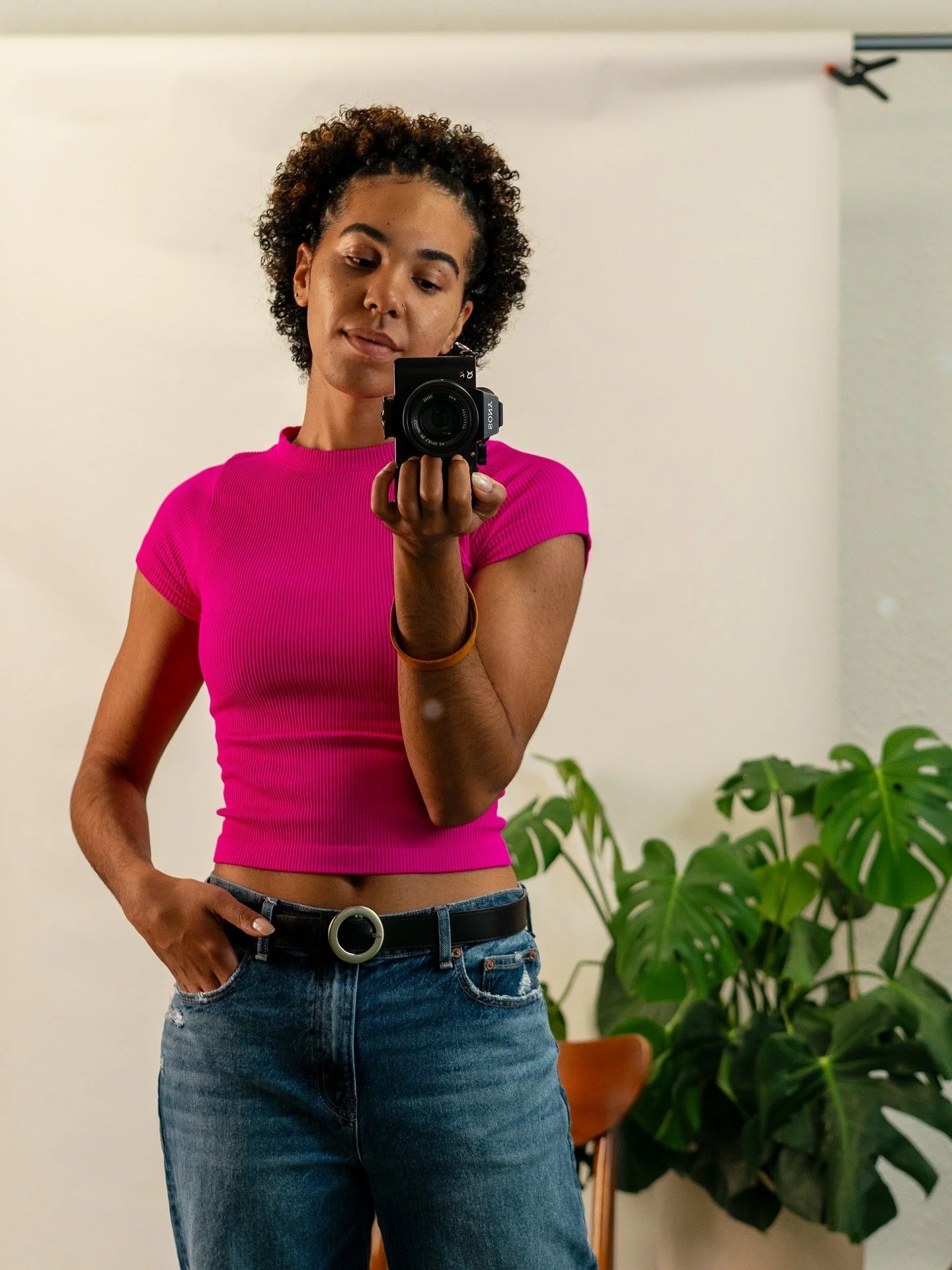 Young woman with curly hair taking a mirror selfie with a camera, wearing a bright pink top and jeans, standing in front of a white wall with a green plant nearby.