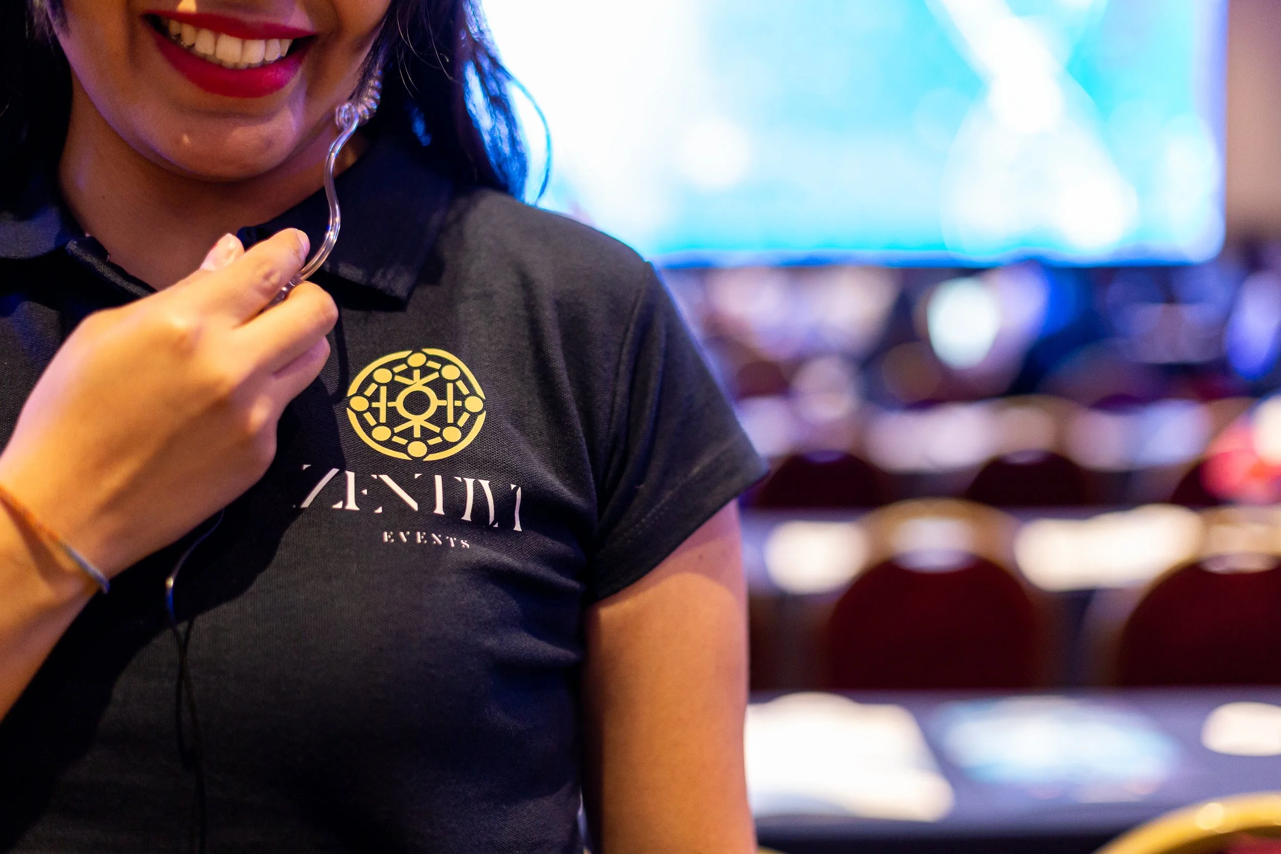Mujer sonriendo con uniforme de evento, sosteniendo un auricular, en un lugar con fondo borroso con pantallas y sillas.