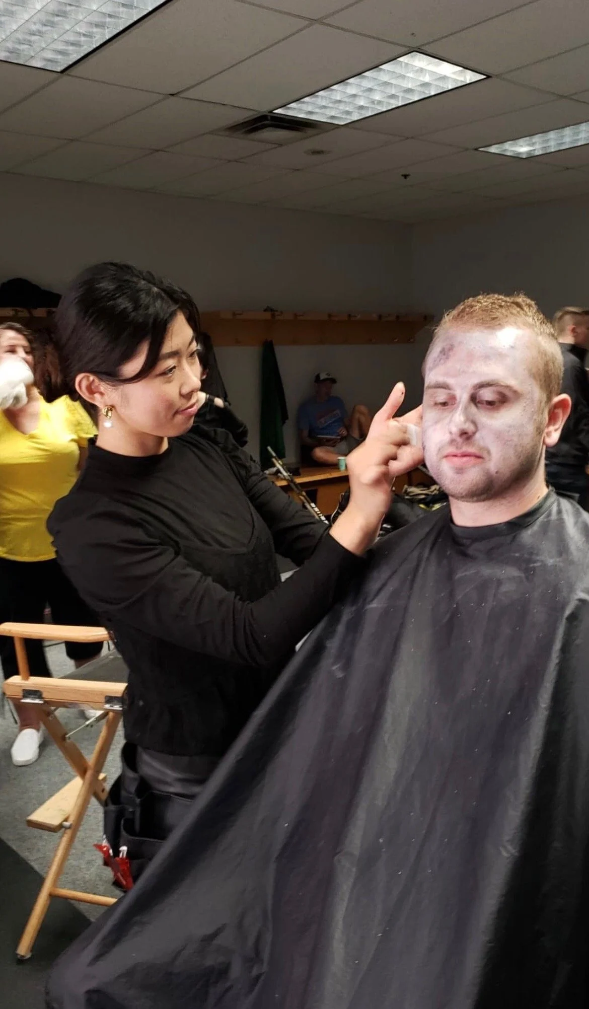 Kana Okada - Experienced Makeup Artist preparing an actor to look like a zombie, with pale makeup and a distressed expression, on a man sitting in makeup chair, in a room with other people and makeup tools.