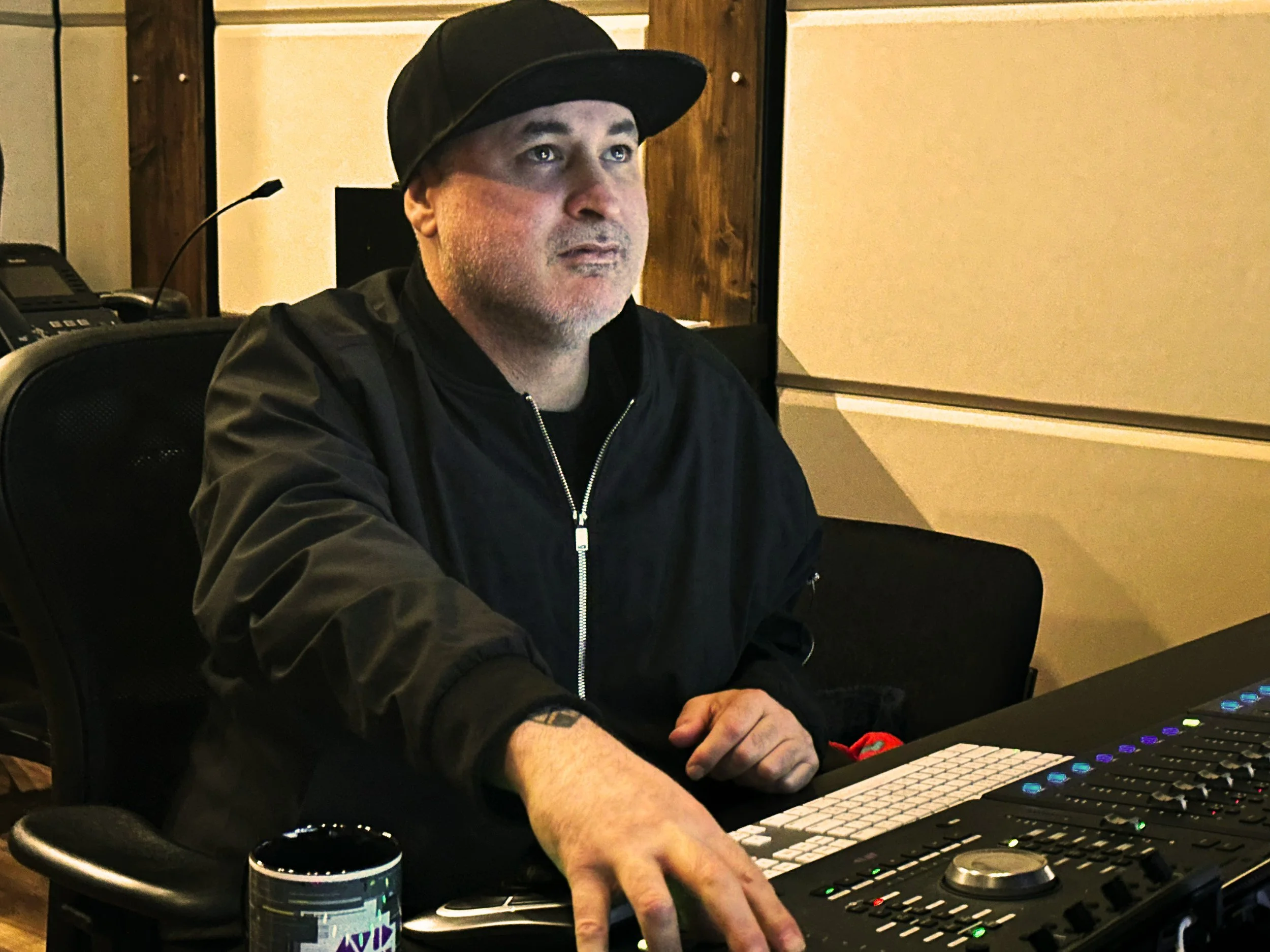 John Franco  – Award-winning Sound Editor – wearing a black cap and black jacket sits at a mixing console in a recording studio, with a coffee mug nearby.