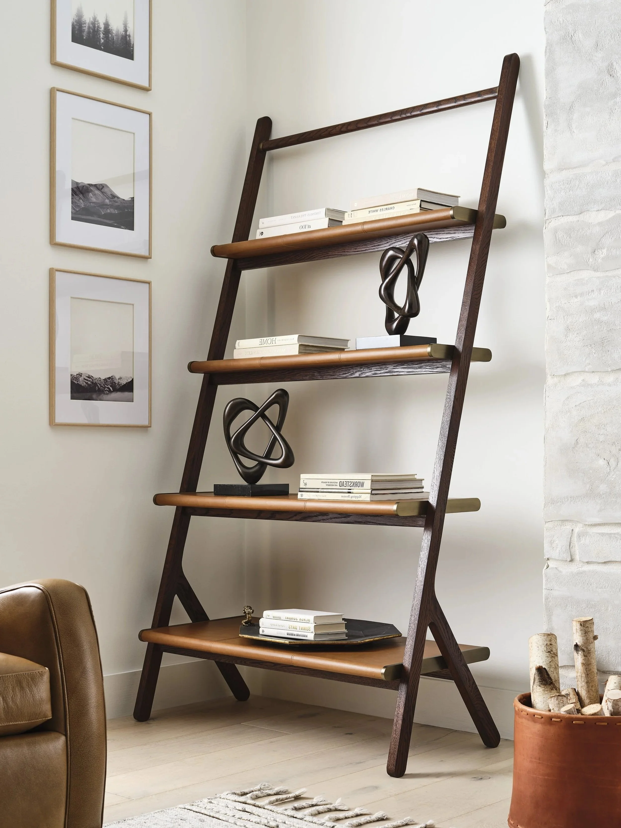 A wooden ladder-style bookshelf with five shelves, decorated with books and black abstract sculptures, against a white wall in a cozy living room.