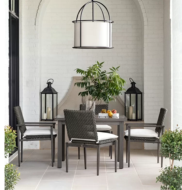 An indoor dining area with a gray table, four wicker chairs with white cushions, a large green potted plant in the center, two black lanterns with candles, and a white brick wall background hanging lamp.
