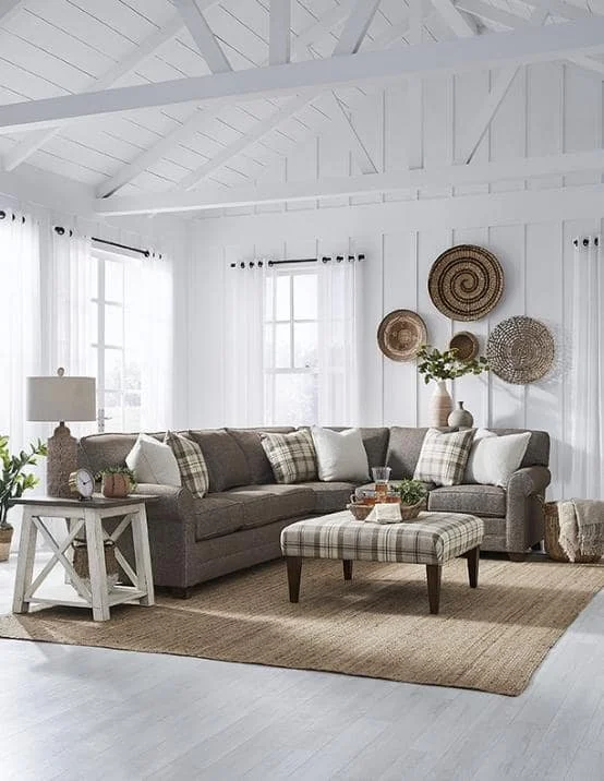 Living room with a gray sectional sofa, plaid and solid pillows, a plaid ottoman, a white side table with a lamp, and wall decorations of woven baskets on white walls with large windows and white curtains.