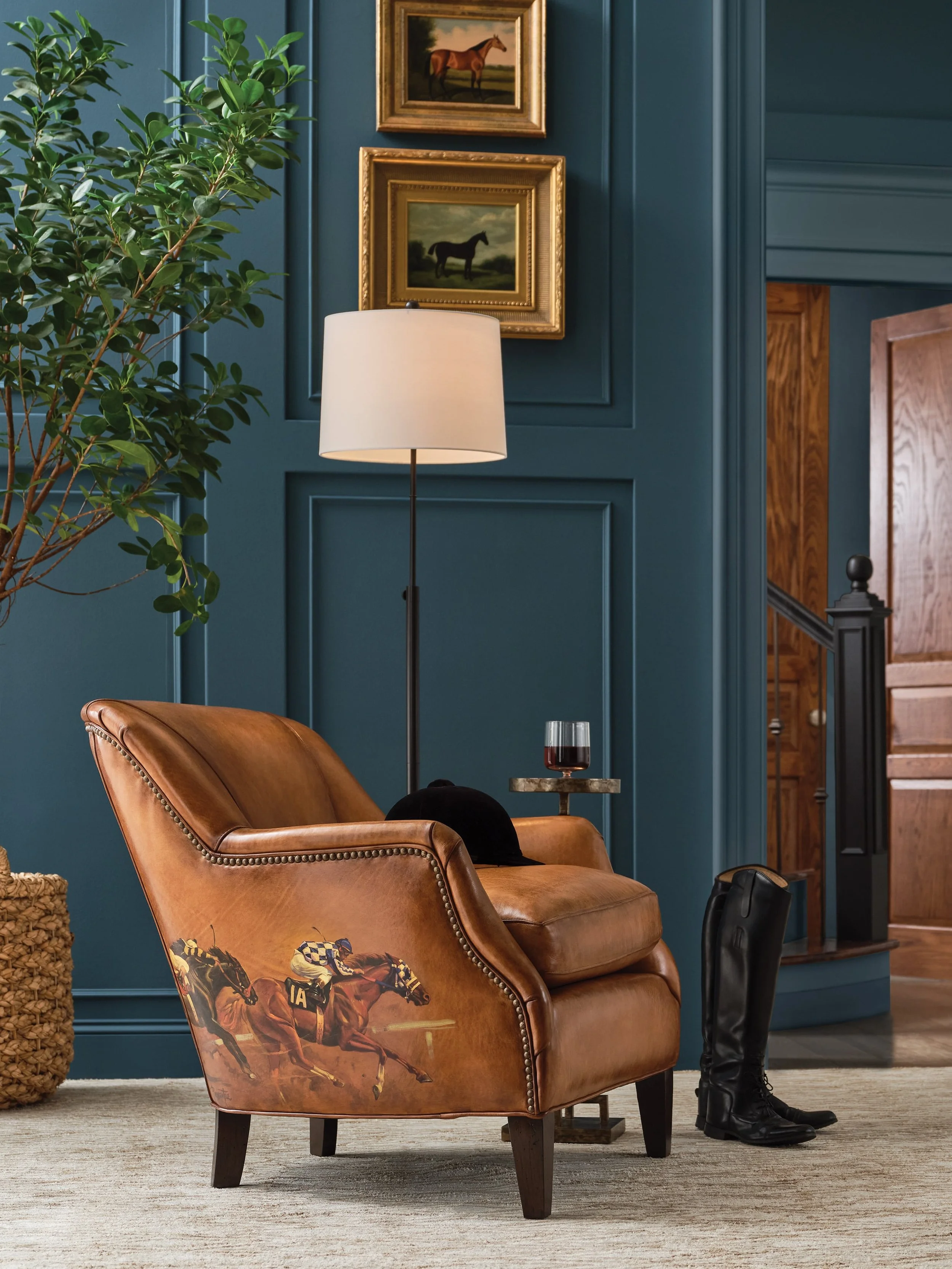 A cozy living room corner with a brown leather armchair featuring painted horse race images, a glass of red wine on a small side table, a black riding boot on the floor, a tall white floor lamp, a large potted plant, and framed horse paintings on a blue wall.
