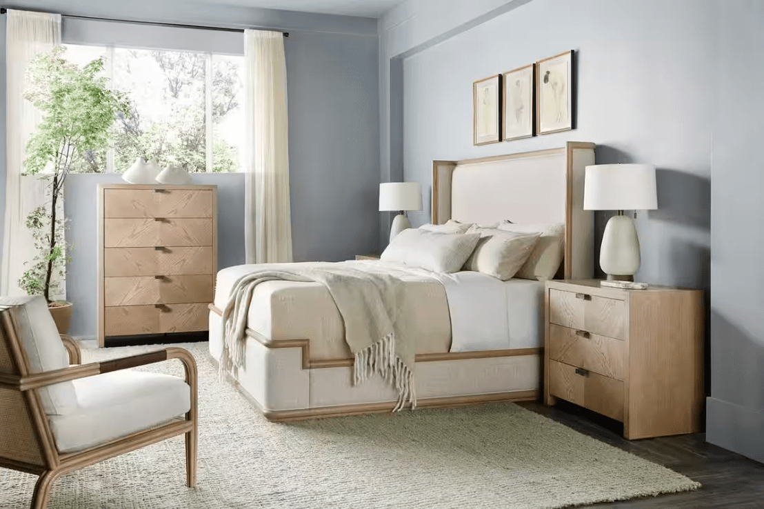 A bedroom with a light blue wall, wooden furniture, a large bed with white bedding, nightstands with lamps, a dresser, a wooden armchair, and a large window with curtains and a view of greenery outside.