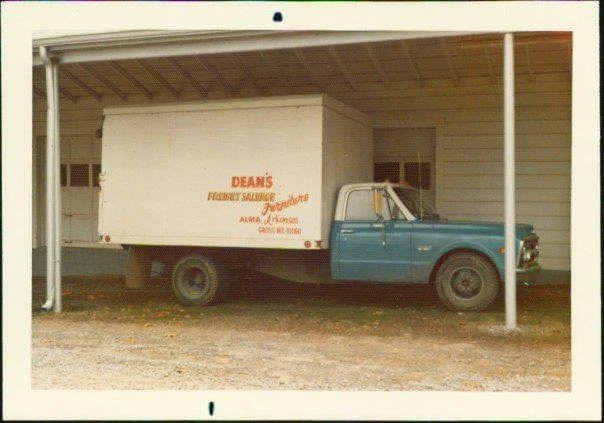A vintage blue and white truck with a large white box on the back, parked under a shelter in front of a white building.