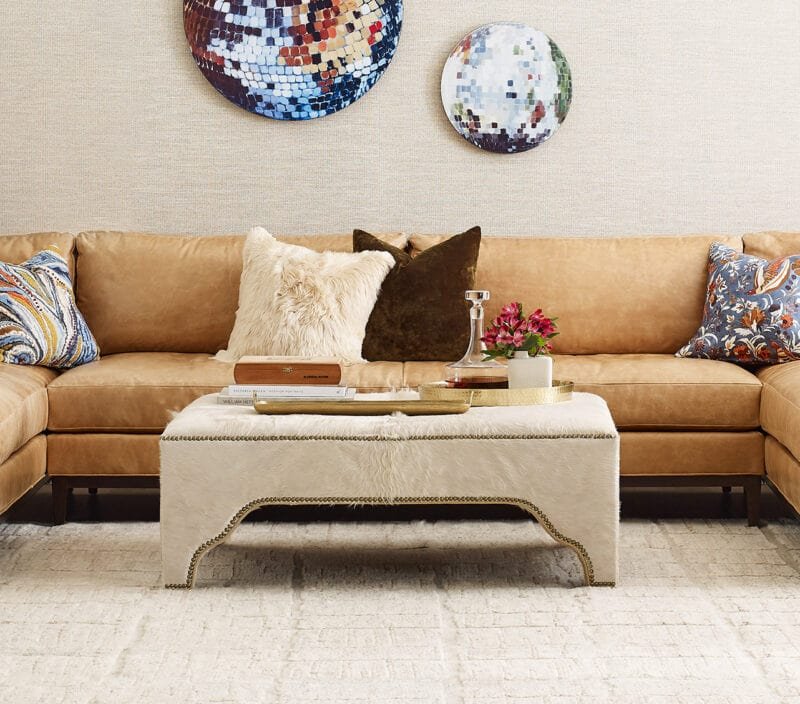 Living room with a beige sectional sofa, decorative pillows, a white coffee table with a tray, books, a glass decanter, and a potted plant; abstract art on the wall.