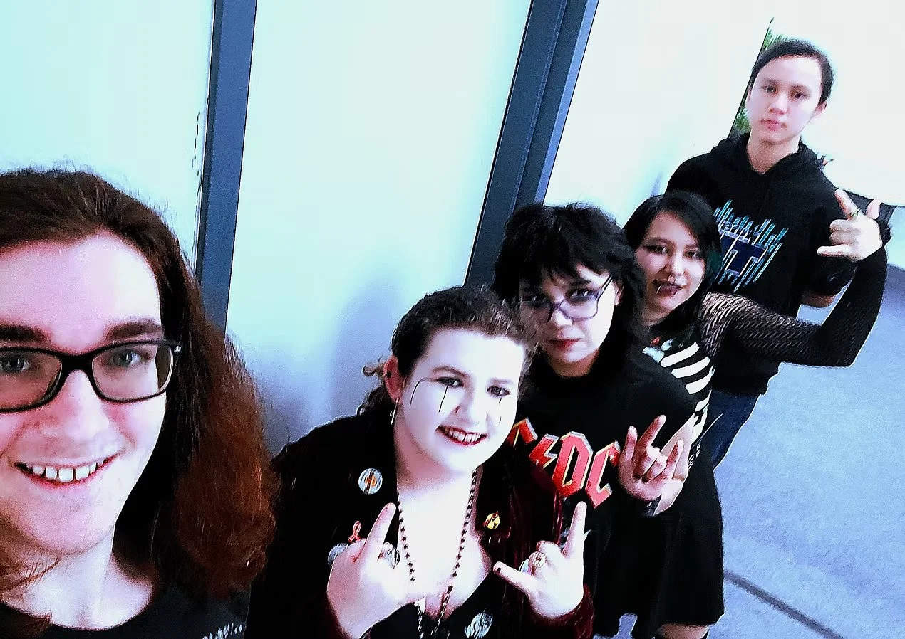 Group of five young women with alternative fashion, some making rock hand signs, posing together indoors.