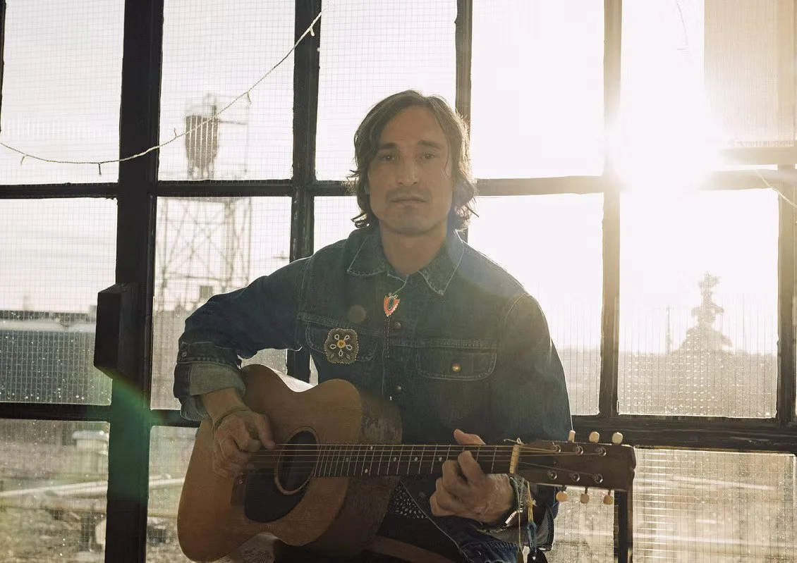 Man playing acoustic guitar in front of a large industrial-style window with sunlight streaming through
