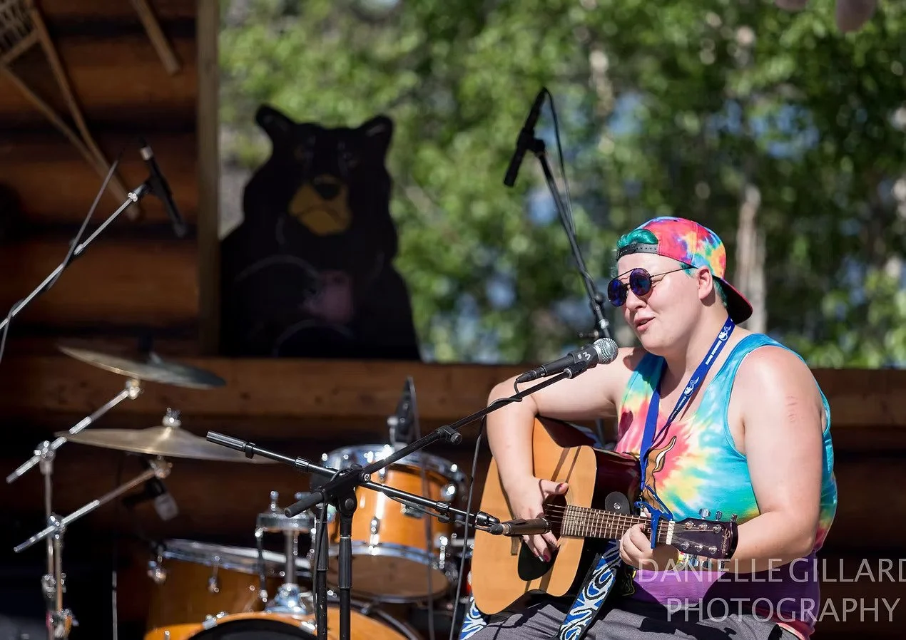 Person wearing a colorful tie-dye tank top, sunglasses, and a tie-dye cap, playing an acoustic guitar and singing into a microphone during an outdoor performance, with a drum set and a bear-shaped black speaker in the background.