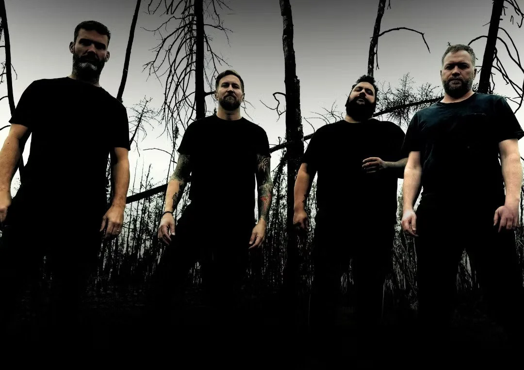 Four men standing in a dark, charred forest with leafless trees and an overcast sky.