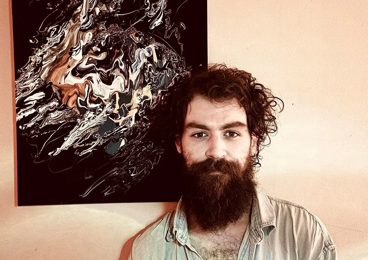A young man with curly hair and a beard standing in front of an abstract black and white painting on a beige wall.