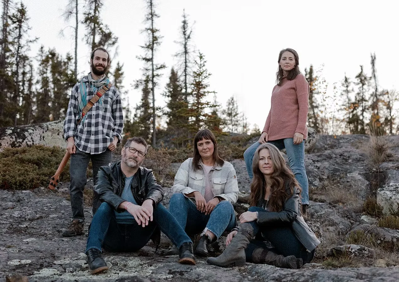 Group of five people outdoors on rocky terrain with trees in the background, some sitting and others standing, dressed in casual clothing.