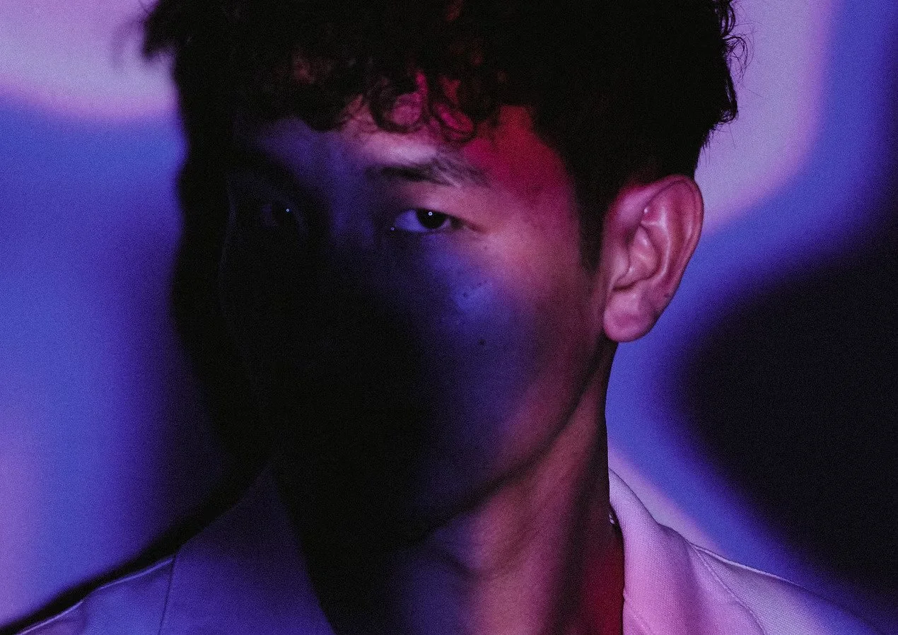 A young man with dark hair and earrings, half of his face in shadow and illuminated by purple and red lighting.
