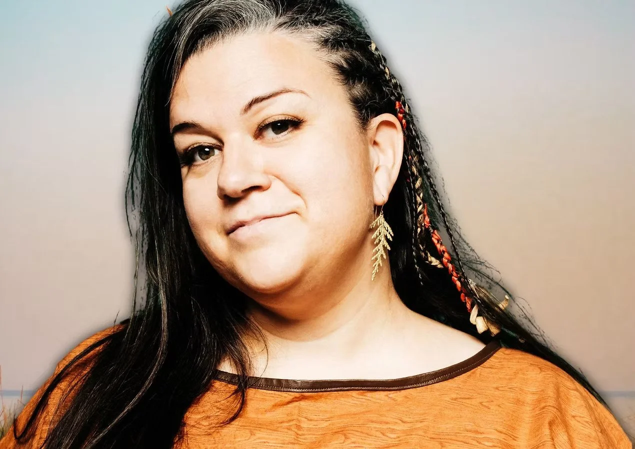 A woman with dark hair and colorful braids, wearing gold leaf earrings and an orange top, standing against a plain background.