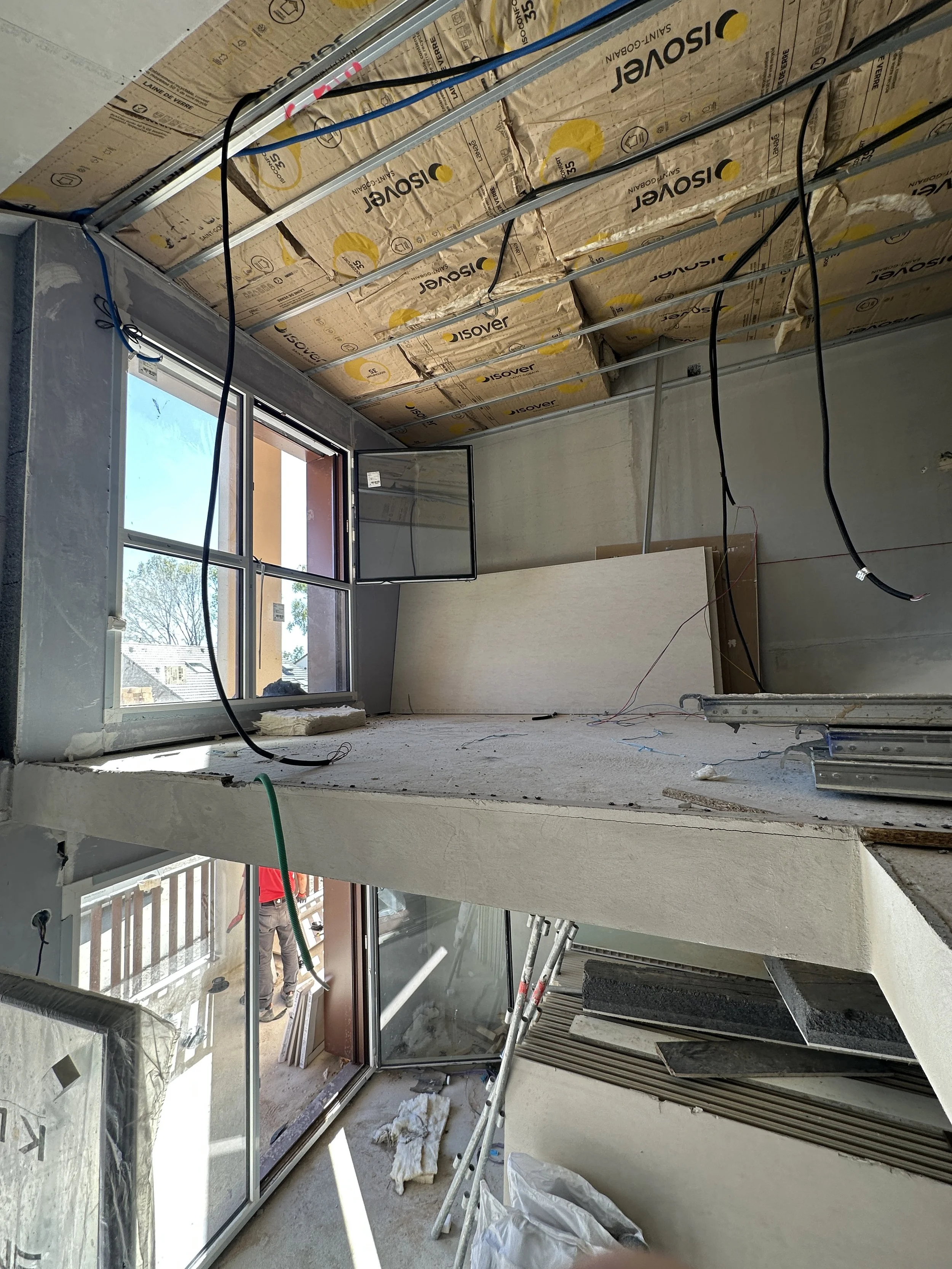 Une pièce en cours de construction avec des fenêtres en verre, un plafond en isolation et des outils de construction tels qu'une échelle, des fils électriques, et des matériaux entreposés.