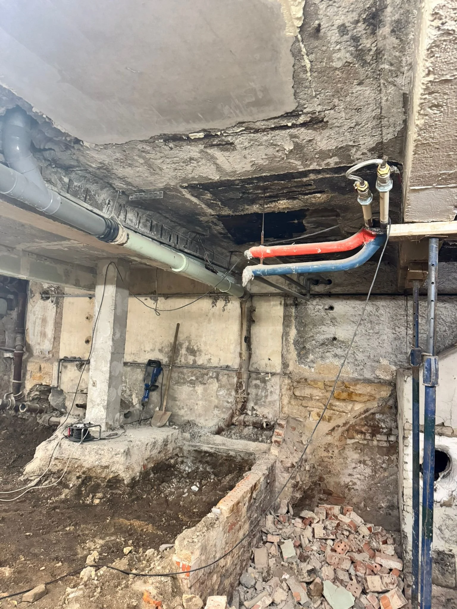 Travaux de rénovation ou de construction intérieure avec murs en cours de démolition, tuyaux et étais en place, débris de briques et de béton au sol.