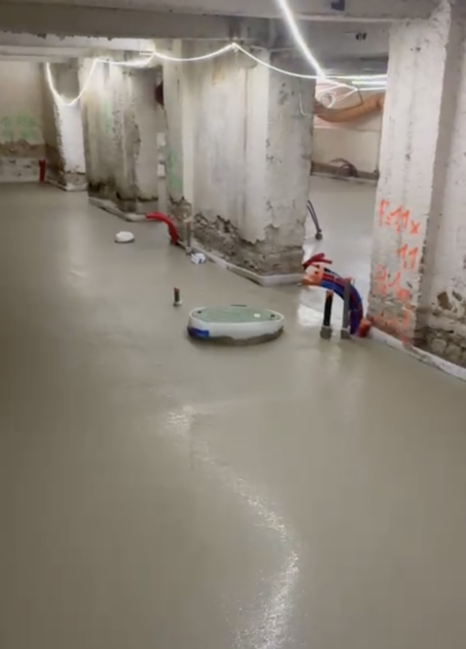 Travaux de rénovation d'un sous-sol avec un sol en béton fraîchement coulé, des tuyaux rouges et noirs visibles, des murs en béton brut avec des débris, des câbles lumineux suspendus, et des marques de peinture orange.