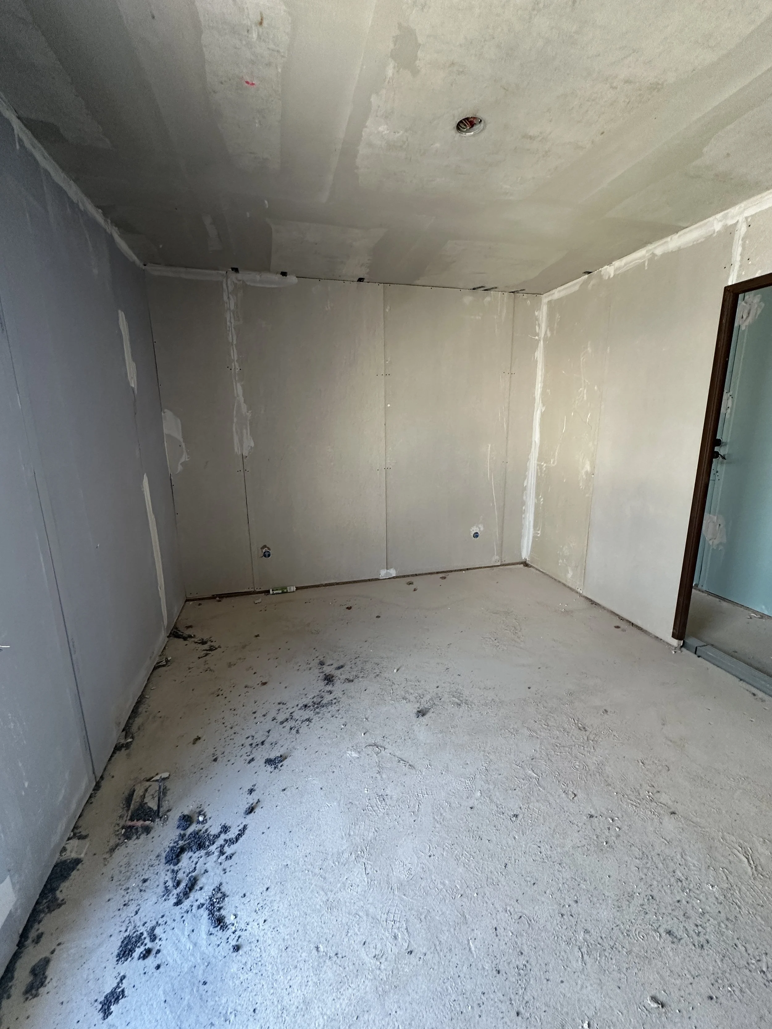 Pièce en construction avec murs en plâtre et sol en béton, il y a des traces de poussière et de débris sur le sol.