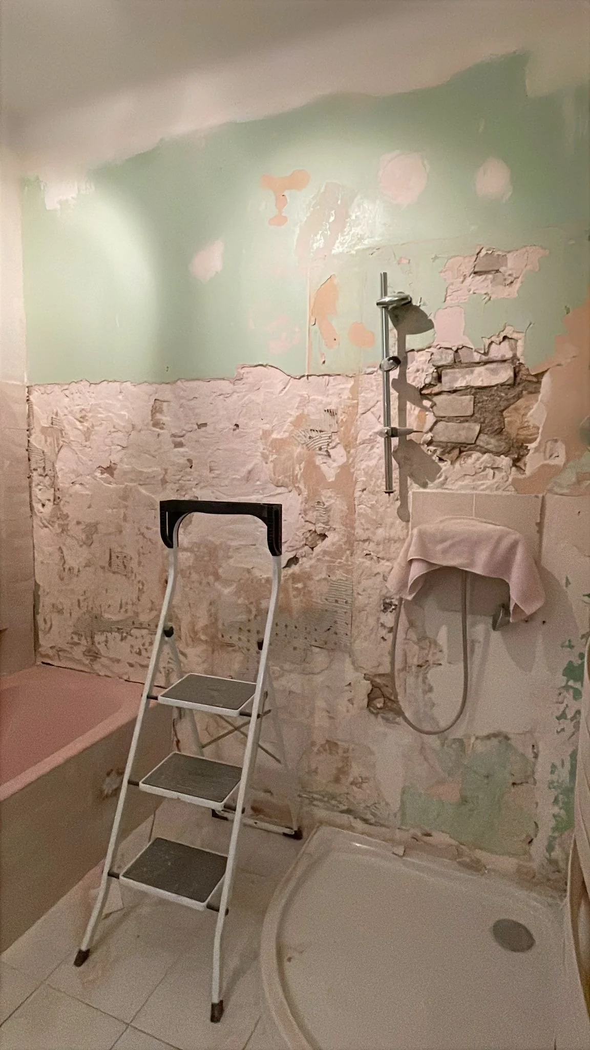 Salle de bain en rénovation avec murs partiellement dégradés, une échelle à trois marches, une douche avec une paroi partiellement installée, et des murs en train d'être réparés.
