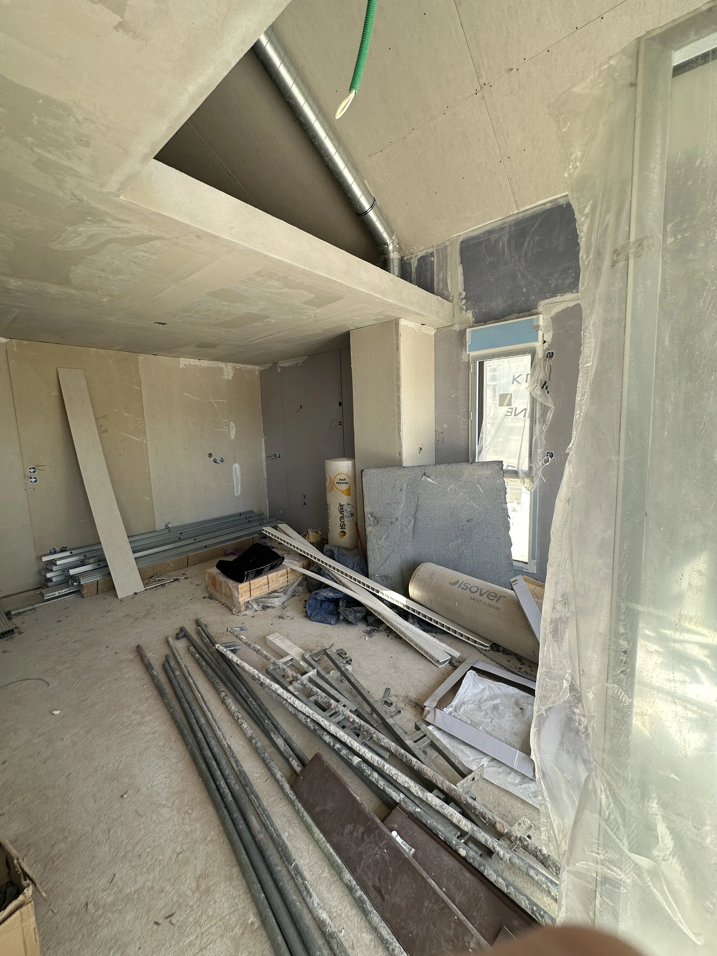 Intérieur d'une pièce en construction avec des outils, des tuyaux, du matériel de chantier, des plaques de plâtre, et un mur inachevé.