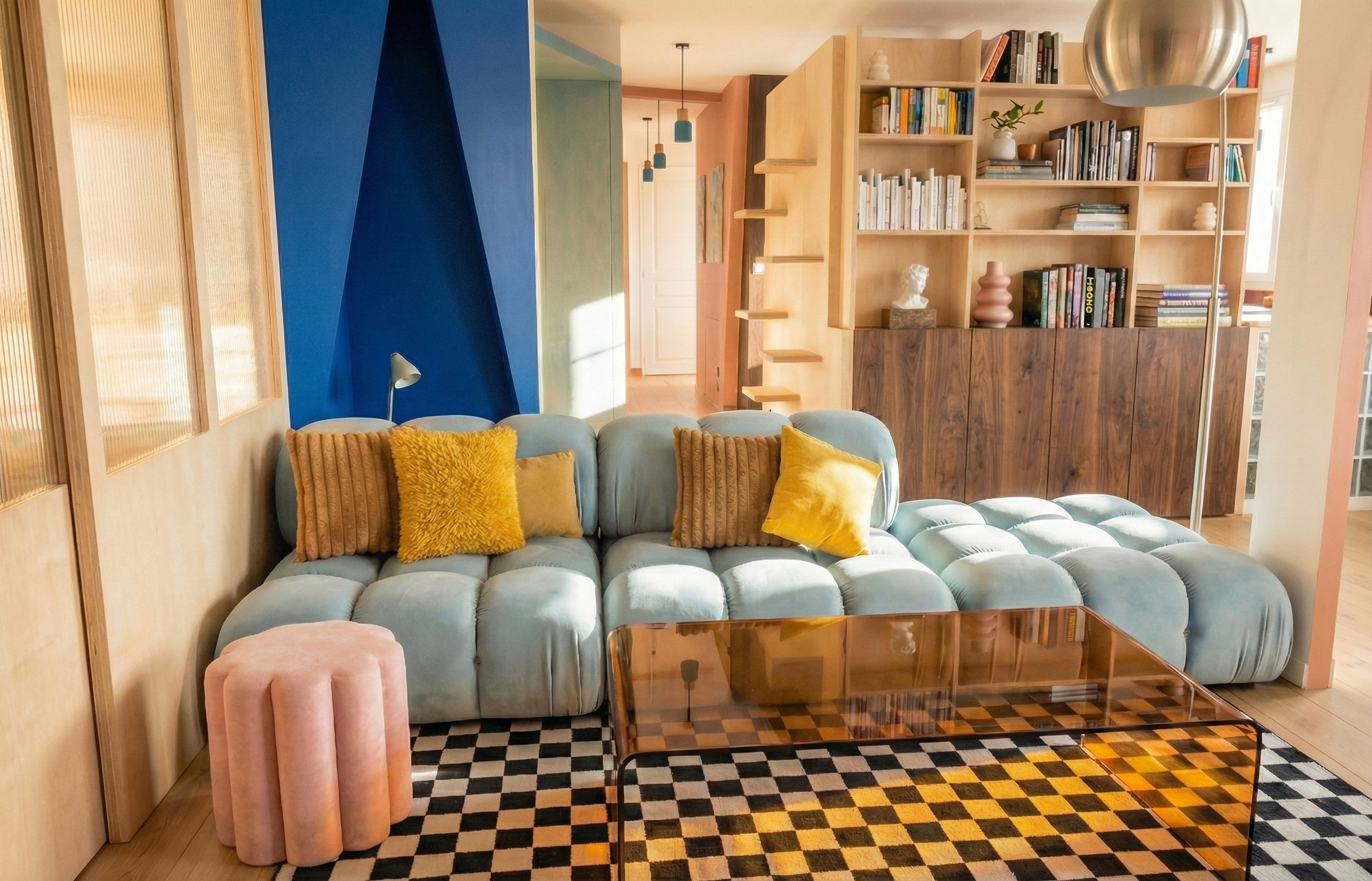 Salon moderne avec canapé bleu, coussins jaunes et bruns, tapis à motif de damier, étagère en bois avec livres et décorations, lampe sur le canapé, lampe sur pied en métal, escalier en bois en arrière-plan, éclairage naturel