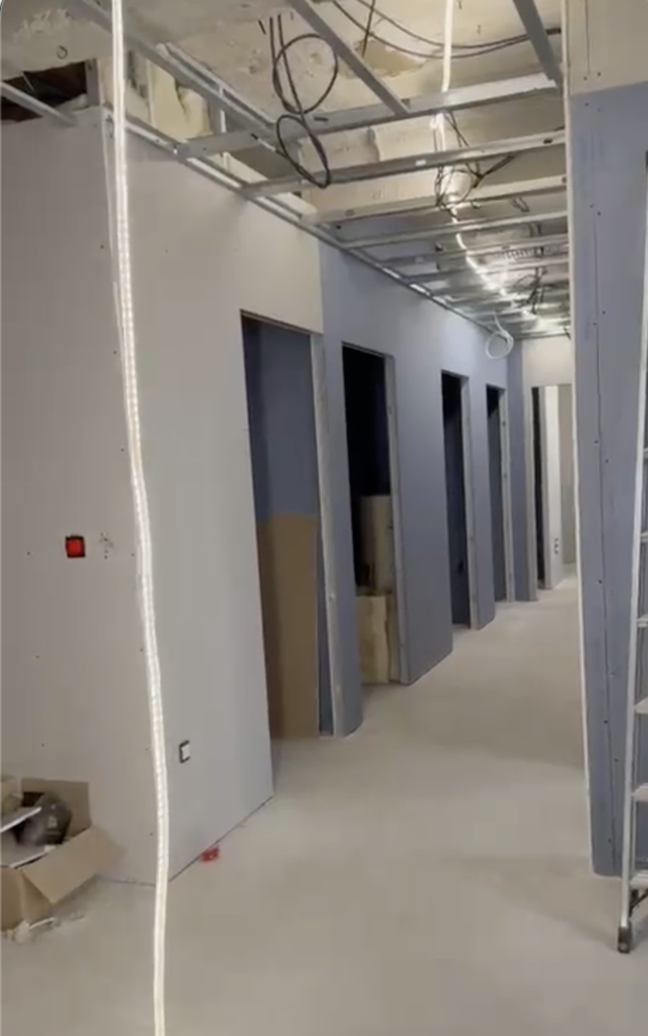 Intérieur d'un bâtiment en construction avec plusieurs portes sans portes installées, murs en plaques de plâtre, câbles électriques apparents au plafond, sol en béton, et une échelle visible à droite.