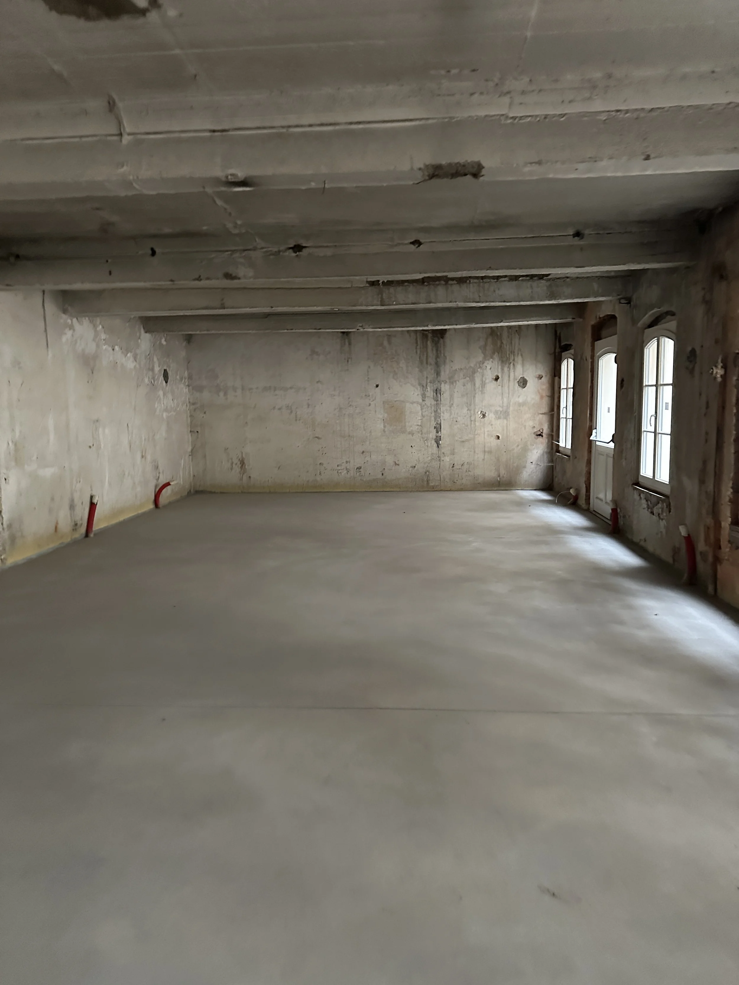 Une pièce en rénovation avec murs en plâtre brut, sols en béton, fenêtres avec cadres en bois et un plafond en béton avec solives apparentes.