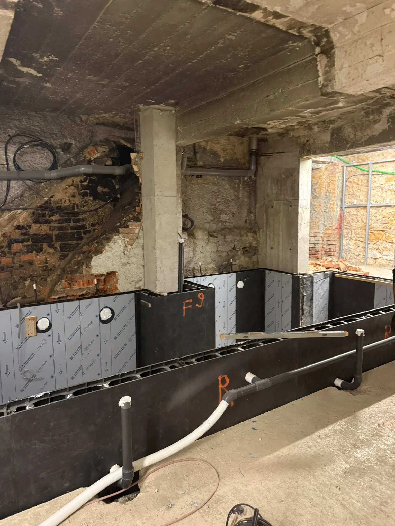 Travaux de construction ou de rénovation avec une cloison en drywall isolé, des tuyaux de plomberie visibles, un mur en brique partiellement démoli et un sol en ciment.