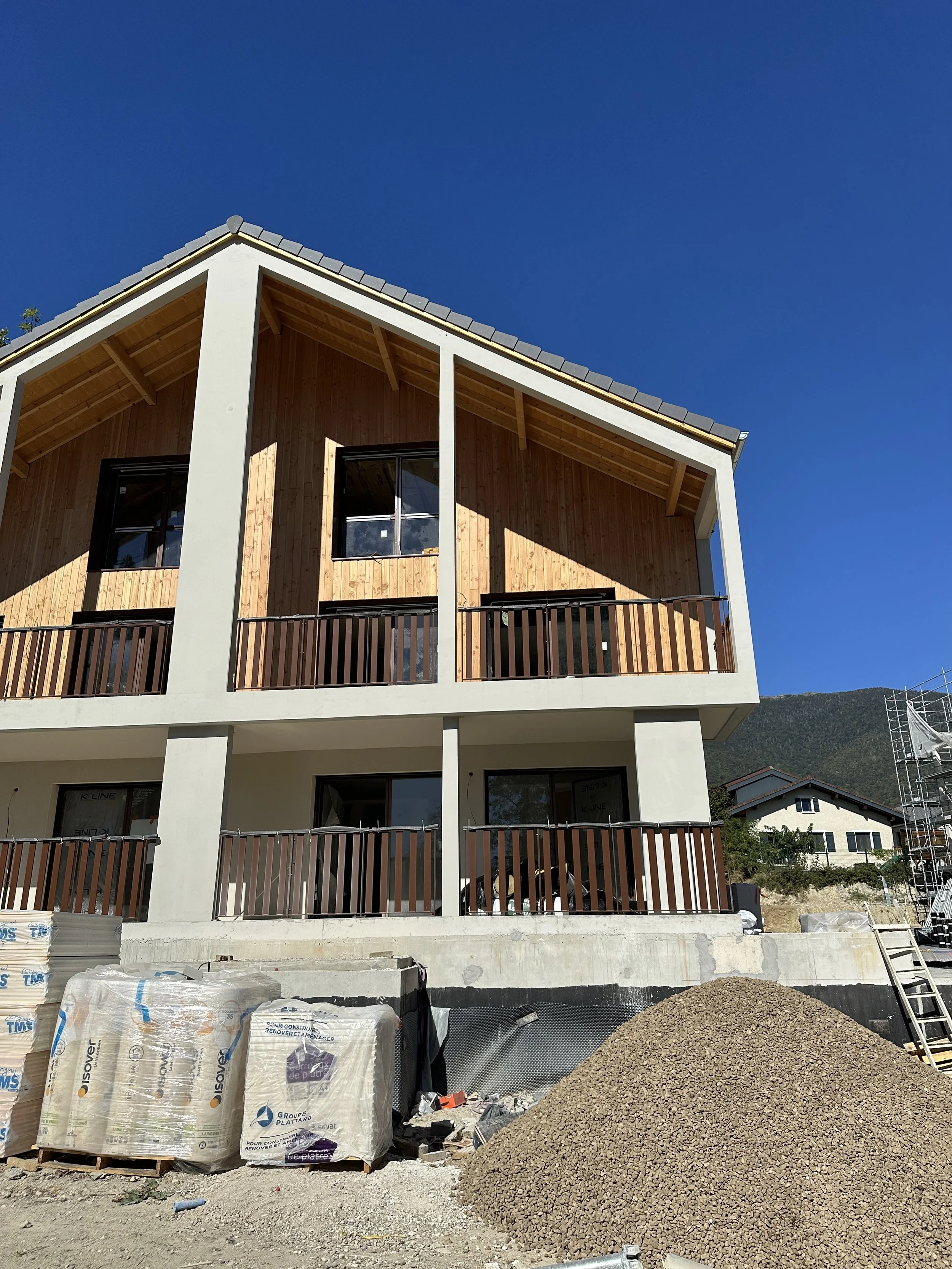 Construction d'un bâtiment résidentiel avec des matériaux de construction en avant-plan, murs en bois et fenêtres installées, ciel clair et montagnes en arrière-plan.