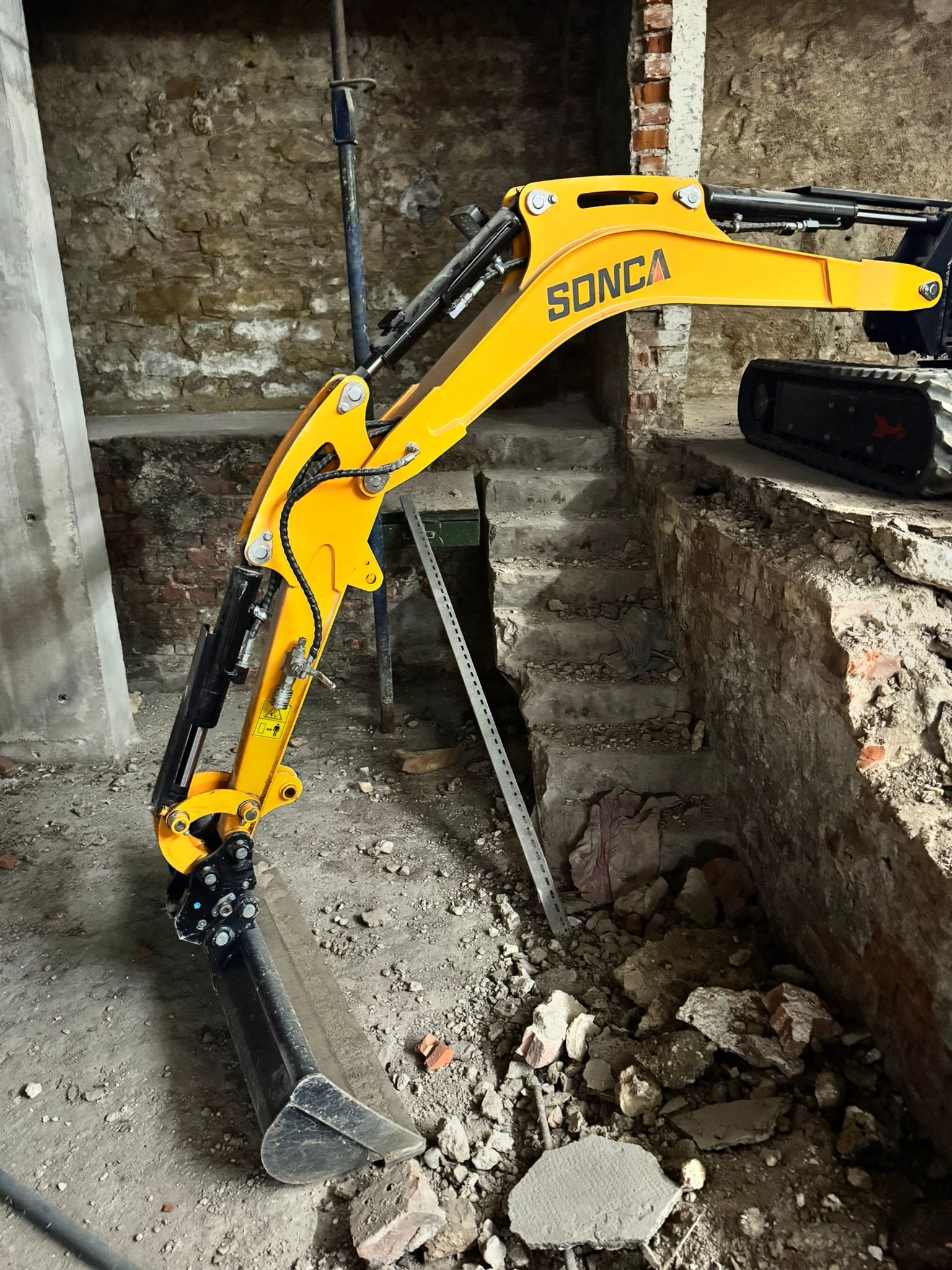 Une excavatrice jaune en plein travail dans un espace en cours de rénovation, avec des murs en pierre et des débris au sol.