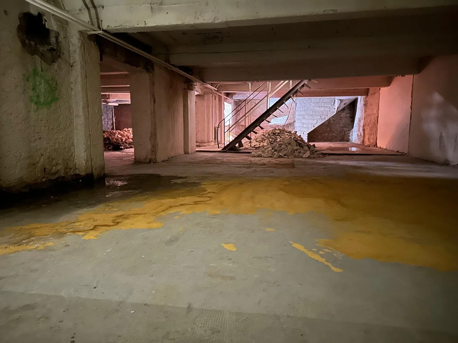 Vue d'un niveau souterrain en construction, avec des piliers en béton, un sol en béton, un escalier métallique en arrière-plan, des débris de construction et des murs en briques.
