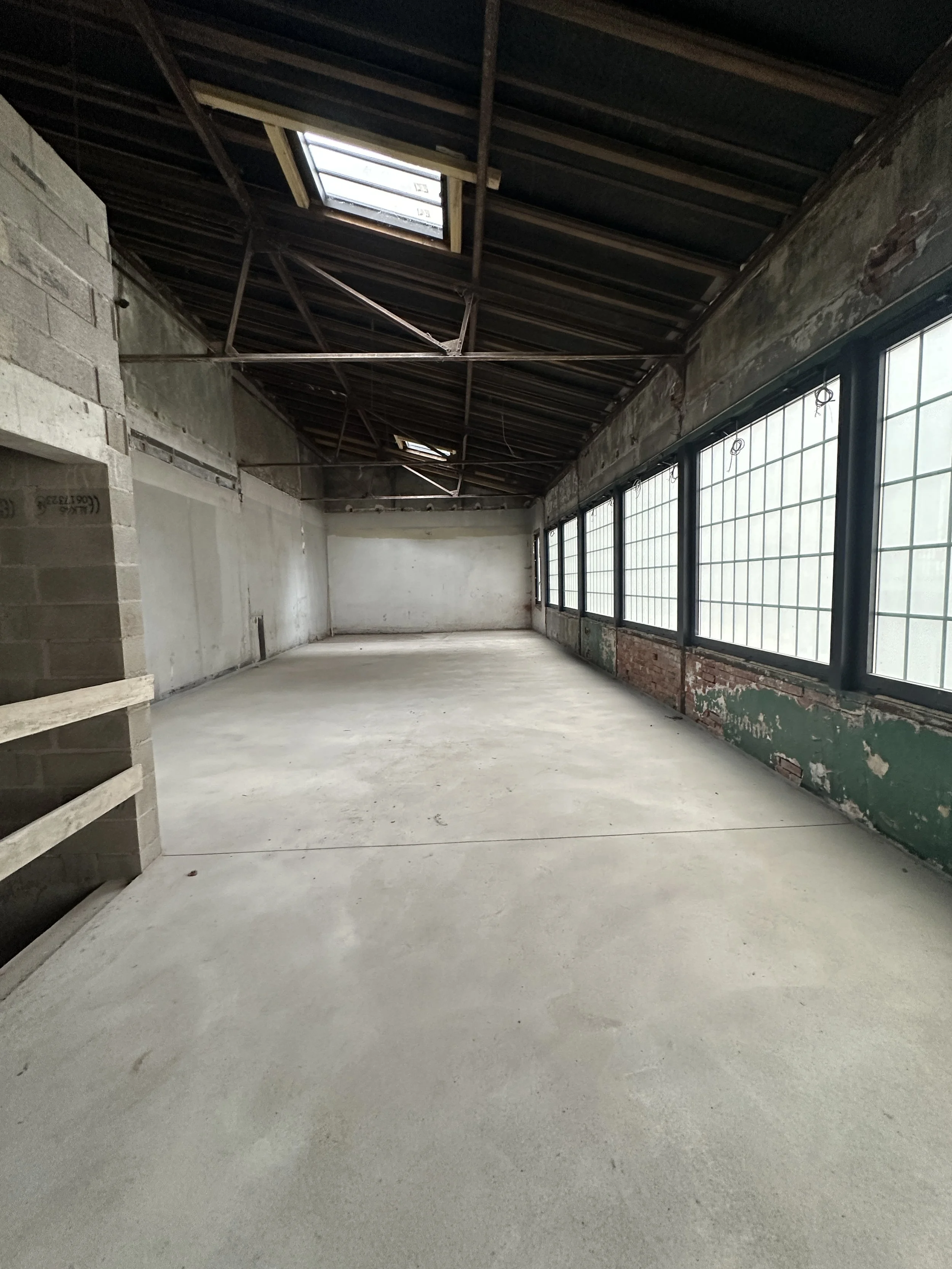 Intérieur d'une pièce en construction avec murs en béton, fenêtres avec barreaux, sol en béton, et un plafond en structure métallique avec des panneaux de toiture. Il y a aussi des fenêtres laissant entrer de la lumière naturelle.