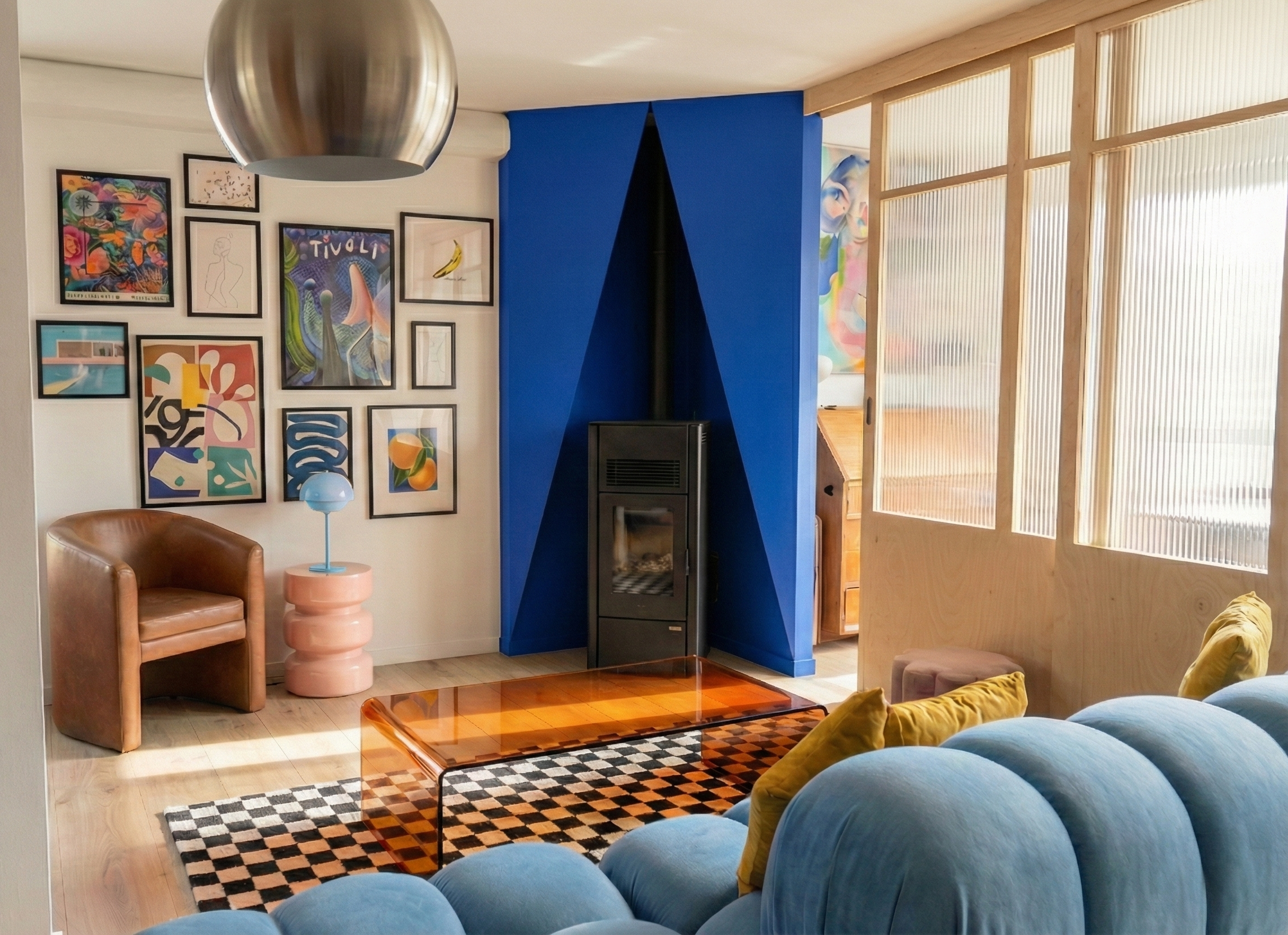 Salon moderne avec mur de tableaux colorés, canapé bleu, fauteuil en cuir marron, cheminée noire encadrée par un mur bleu, table en plastique orange, lampe bleue sur table rose, tapis à carreaux en noir et blanc, divisoires en bois avec panneaux en plastique translucide.