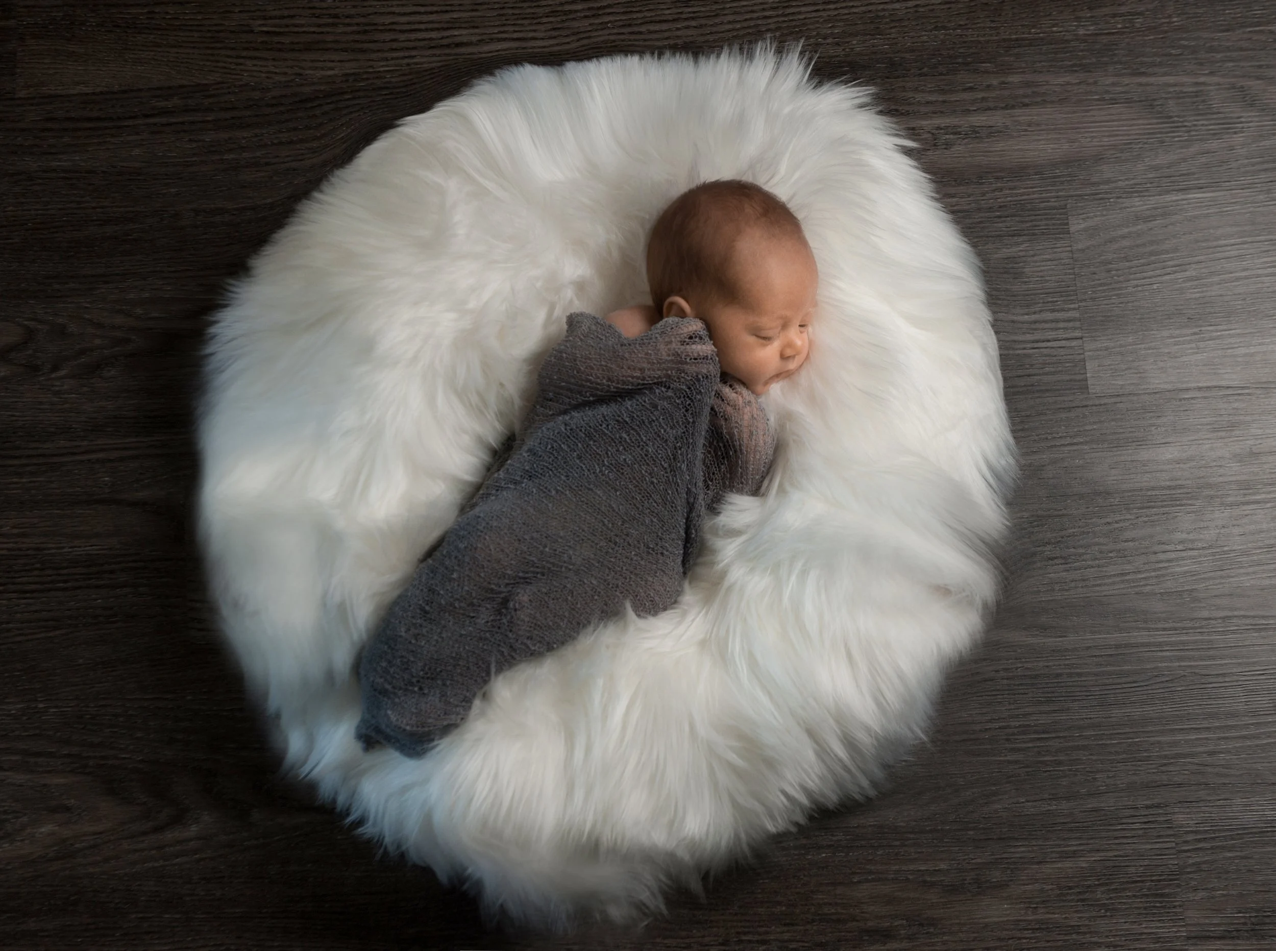 A newborn baby lying on a fluffy white fur blanket on a dark wooden floor, wearing a gray knit outfit and sleeping.