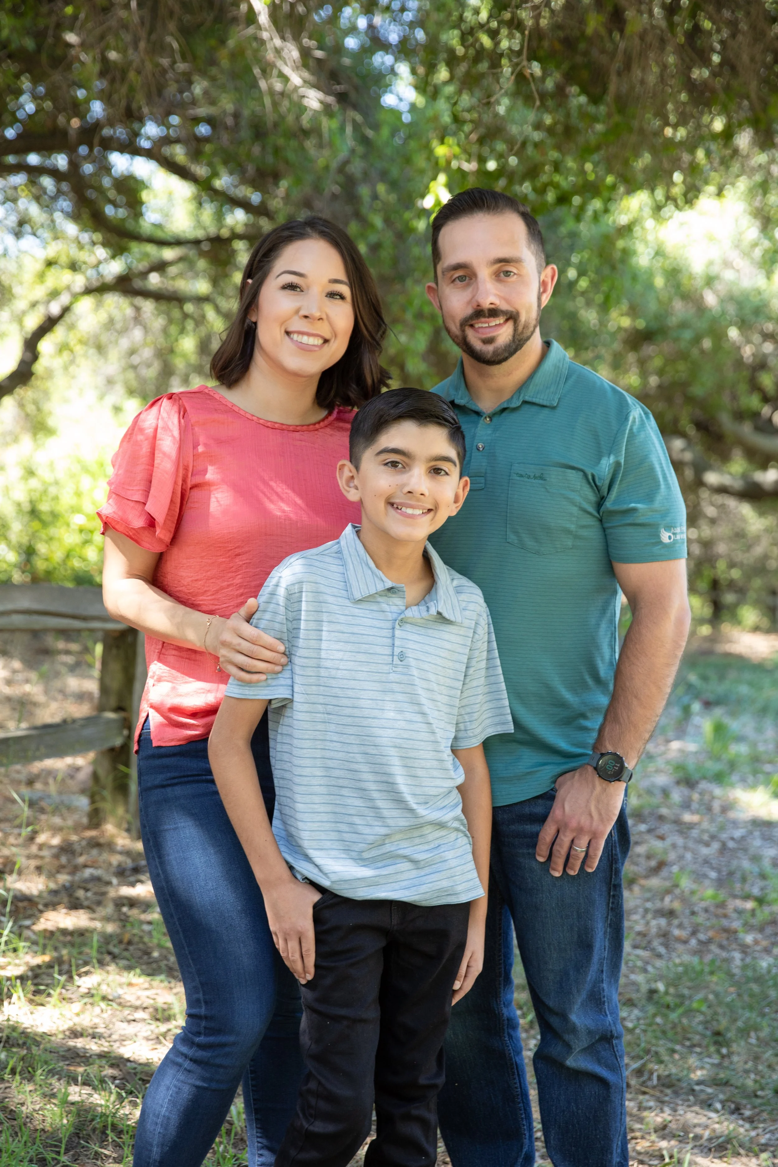 A family of three standing outdoors in a park-like setting with trees, smiling at the camera.