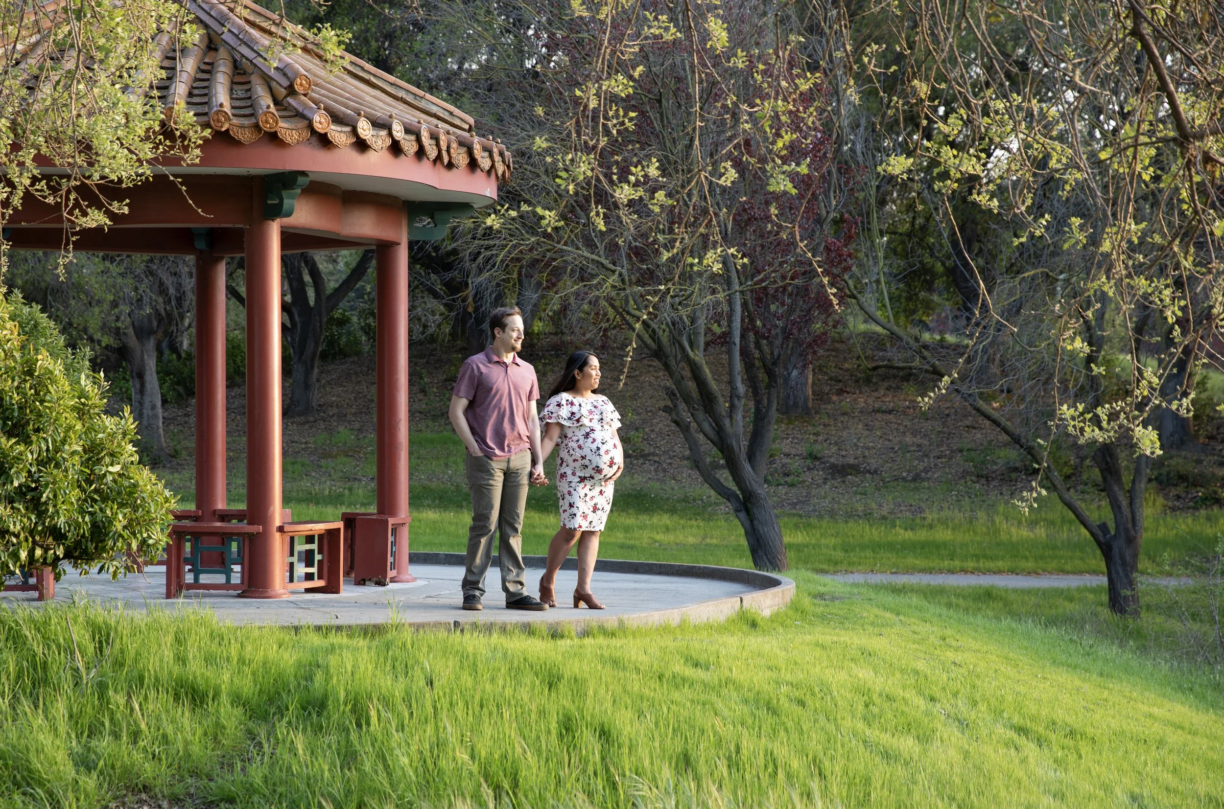A couple, one pregnant woman and a man, standing hand-in-hand on a circular park pavilion, surrounded by green grass and trees with spring leaves.