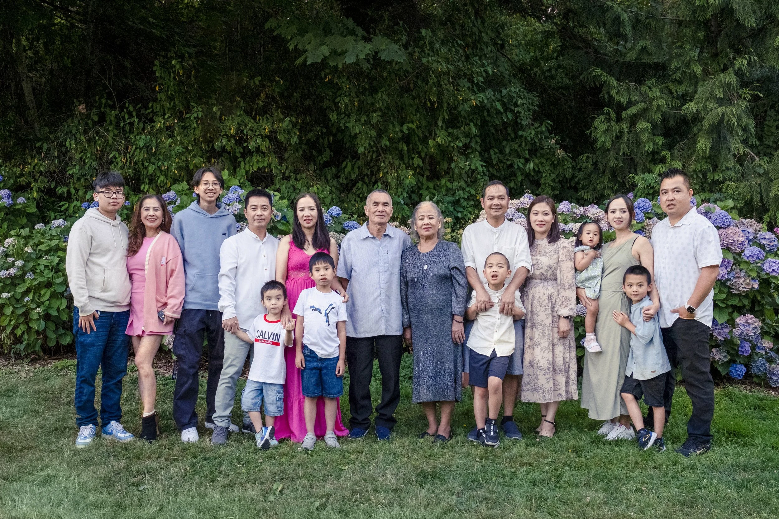 A large family group standing outdoors on grass with a lush green hedge and purple hydrangea flowers in the background, posing for a photo.