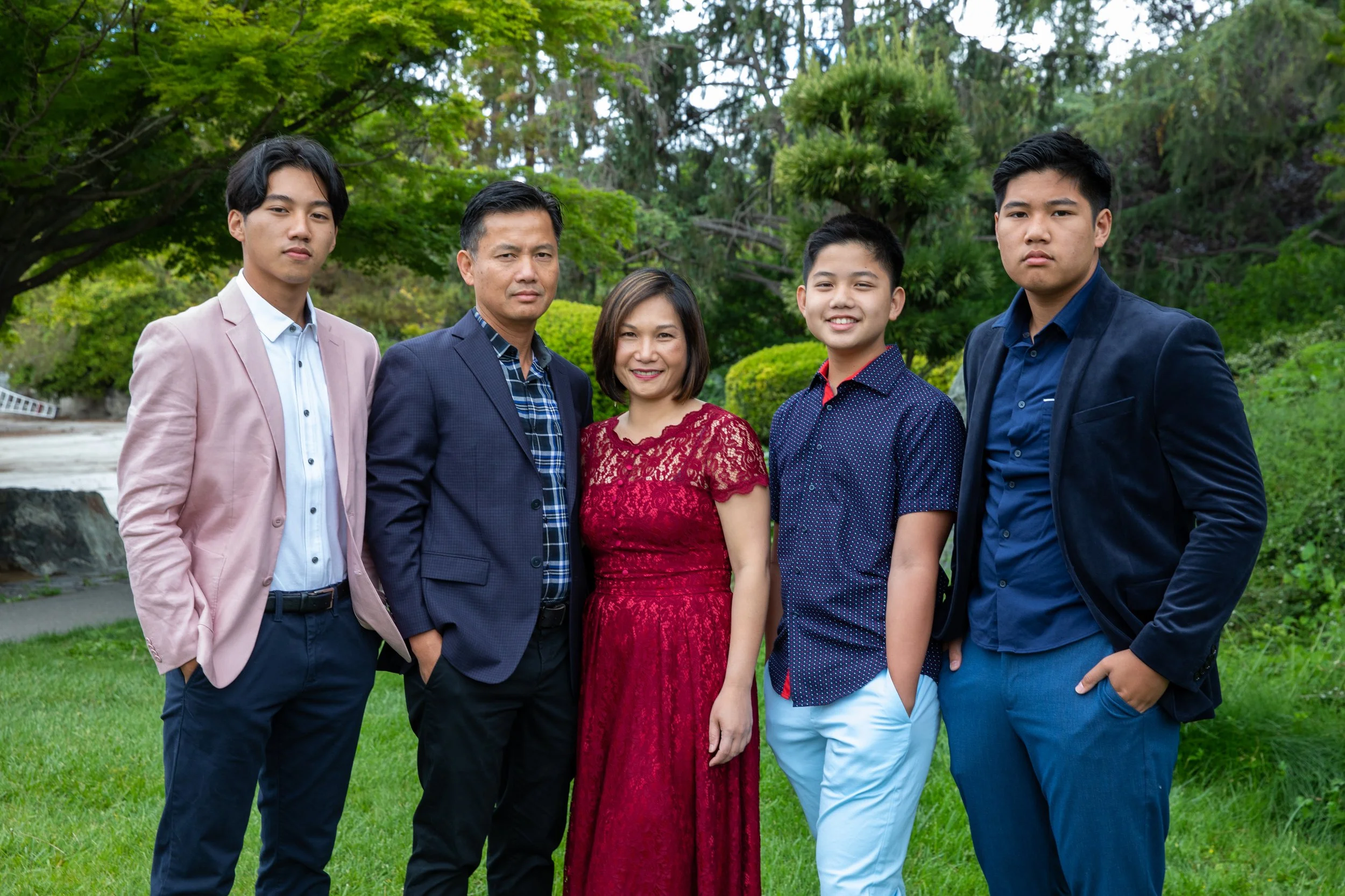 A family of six standing outdoors on a grassy area with trees in the background. The group includes three adult men and women, and two teenage boys. They are dressed in formal and semi-formal clothing and are standing close together, smiling at the c
